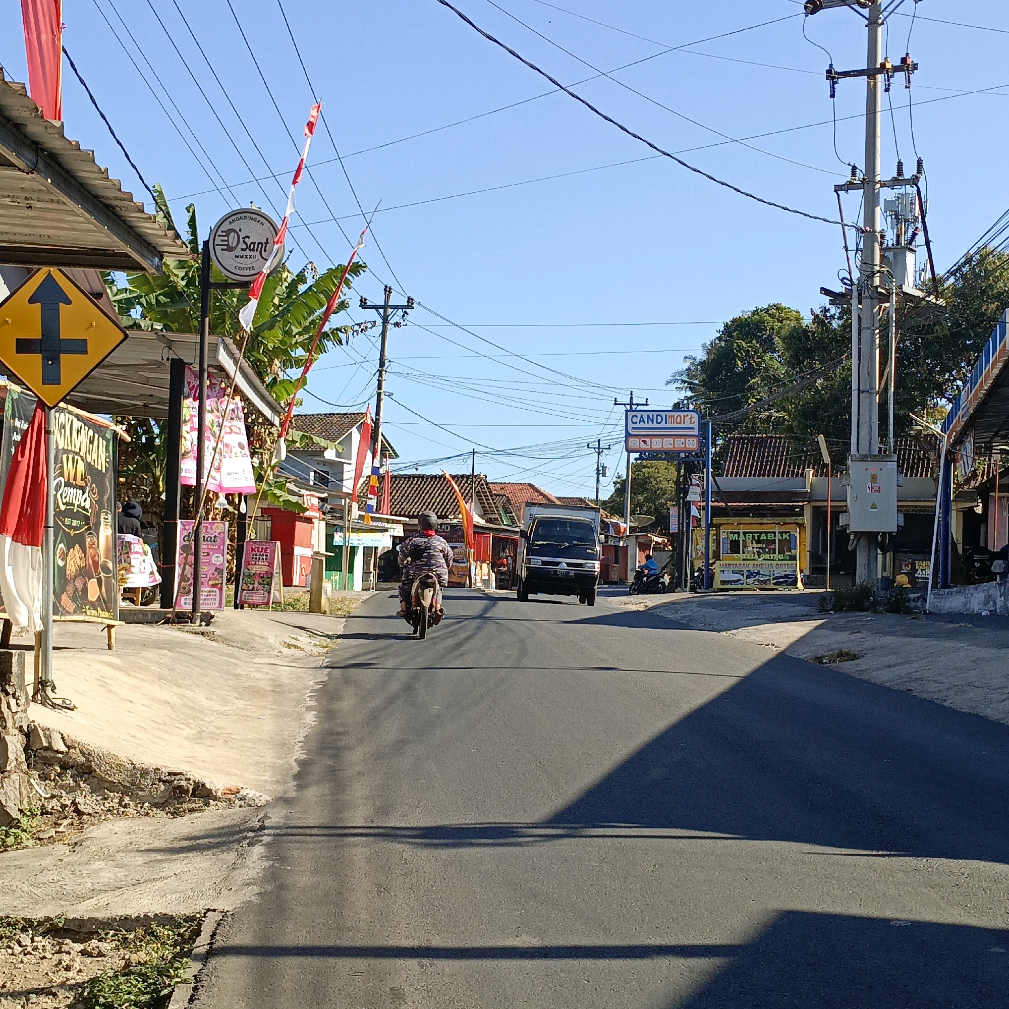 The image depicts a street scene in Candirejo, Indonesia, under a clear blue sky. A paved road runs through the center, with a black van and a motorcycle visible on it. The motorcycle, ridden by a person wearing a camouflage-patterned shirt and a helmet, is in the foreground, traveling away from the camera. The van is further down the road, also moving away.
Along the left side of the road, a row of buildings with corrugated metal roofs are visible. One building has a sign that reads "ANGKRINGAN D'Sant COFFEE". A yellow diamond-shaped traffic sign with a black arrow indicating a straight path and a left turn is prominently displayed. Red and white Indonesian flags are tied to poles and draped from structures. Various banners and signs advertising food and drinks are also present, including one that says "MARTABAK AMELIA ORTEGA".
On the right side of the road, more buildings with red-tiled roofs are visible. A prominent blue sign reads "CANDImart". A utility pole with electrical equipment and transformers stands near the buildings. Trees with lush green foliage are scattered throughout the scene, particularly behind the buildings. The ground along the roadside consists of a dirt path and some scattered debris. The overall impression is of a bustling village street on a sunny day.