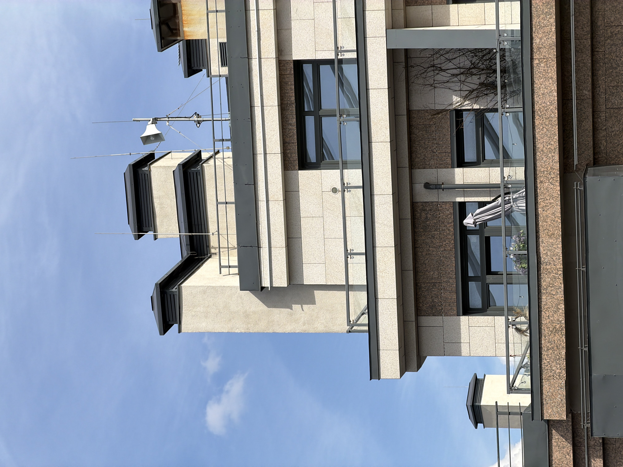 This image captures a detailed section of a modern building facade in Saint Petersburg, Russia, set against a clear blue sky with sparse white clouds. The structure features multiple levels with a textured stone cladding, primarily composed of light beige/cream rectangular tiles and darker brown, possibly granite, sections.
The uppermost visible section of the building includes a flat rooftop perimetered by dark metal railings. From this roof, several large, rectangular, chimney-like structures with dark gray louvered caps project upwards; one cap on the far right exhibits visible rust. Multiple slender metallic rods, likely lightning conductors or communication antennas, extend vertically from the roofline. Centrally positioned on the rooftop is a white, horn-shaped loudspeaker or siren mounted on a metal pole, secured with multiple guy wires, and featuring additional small antenna elements.
Below the rooftop level, a floor with dark-framed, multi-pane windows is visible, fronted by a balcony with glass and metal railings. Further down, a lower balcony is partially visible, also equipped with glass and metal railings. This lower balcony displays a folded grey patio umbrella, a small light-colored object (possibly a pet or decorative item) near the railing on the left, and potted plants with purple flowers. A vertical grey downspout is affixed to the building's facade on this lower level. Sunlight creates strong shadows across the building's surfaces, emphasizing its architectural details. No individuals are present in the depicted scene.
