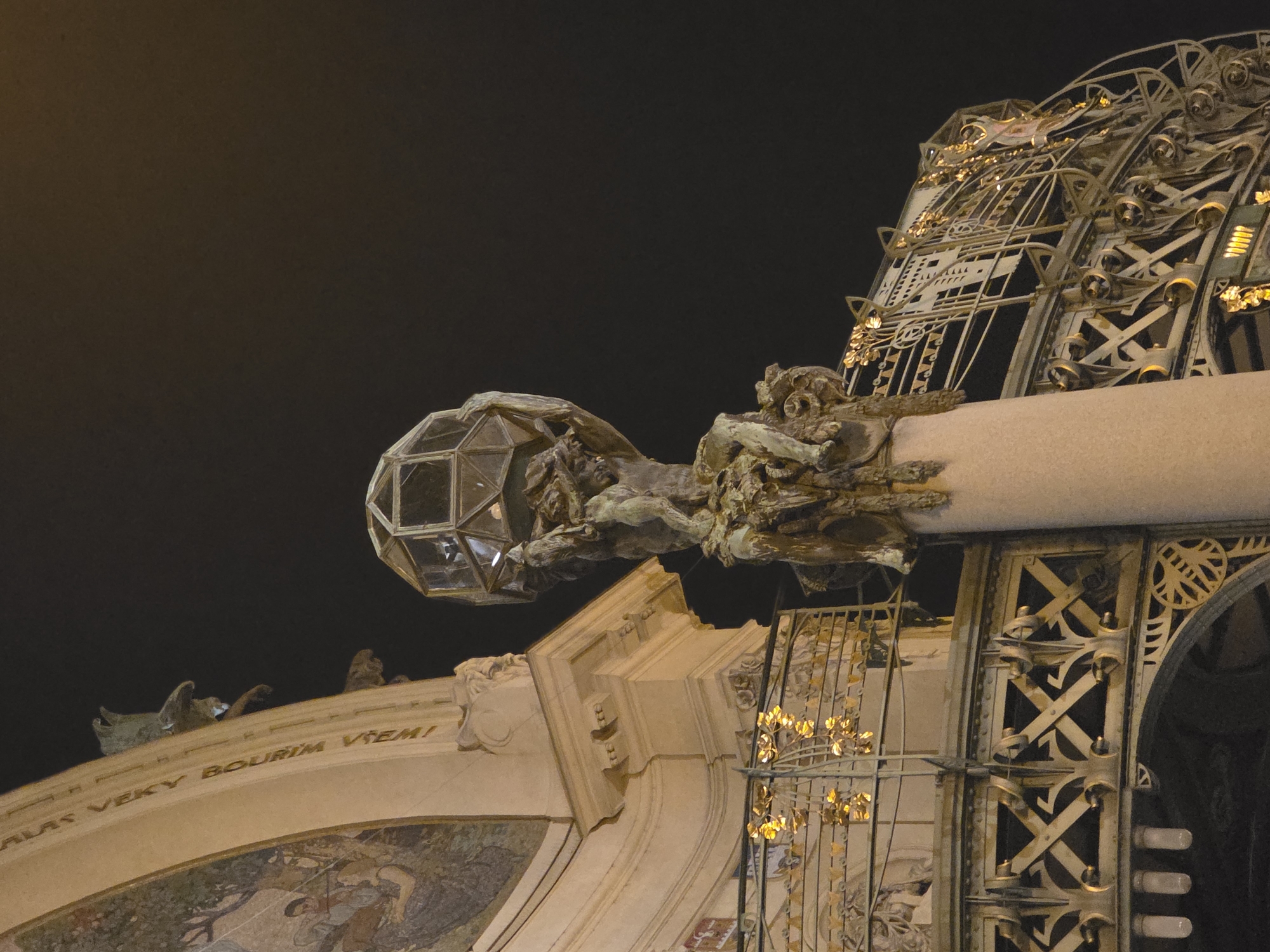 This image captures a detail of an ornate building facade at night, characterized by intricate design and illumination. The dominant feature is a large, elaborate metallic structure, possibly a gate or decorative railing, adorned with golden accents and geometric patterns. This metalwork appears to be illuminated, casting a warm glow.
To the left, a spherical, multi-faceted glass lantern is suspended, held aloft by a complex, aged metal sculpture that resembles a gargoyle or mythological figure. This sculptural element is rich in detail, showing texture and form suggestive of bronze or weathered copper.
Below these features, a light-colored, curved stone archway is visible. Along its upper edge, an inscription is carved, which reads: "HLAS VEKY BOURIM VTEM!". Below the arch, a portion of a colorful mosaic or fresco is discernible, depicting figures and foliage in an artistic style.
The overall impression is one of historical or Art Nouveau architecture, showcasing craftsmanship and decorative artistry. The dark sky indicates nighttime, with artificial lighting highlighting the architectural details and creating a dramatic effect.