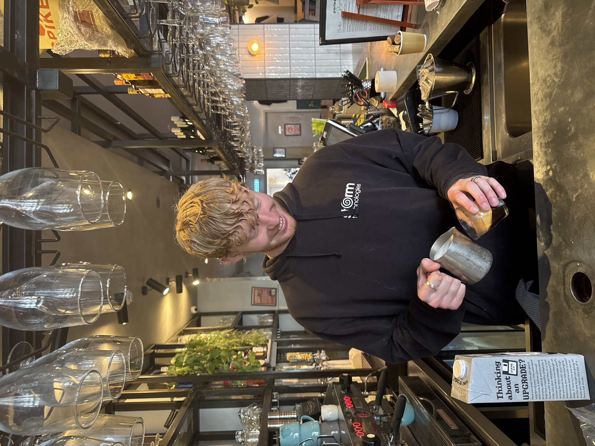 A young, fair-skinned man with blond curly hair is preparing a drink behind a bar. He is wearing a black hoodie with a company logo on the right breast. The logo appears to contain the word "Form" and the word "technologies" underneath. The man is smiling and is in the process of pouring liquid from a small glass container into a metal pitcher. He is wearing a ring on one of his fingers.
The scene is indoors, likely a cafe or bar in London, United Kingdom. The setting includes bar equipment such as an espresso machine and a stainless steel sink. Shelves of wine and glassware are visible in the background, along with green plants. There is a milk carton on the counter bearing the text "Thinking about an UPGRADE?" and other promotional text. The bar area is well lit with natural and artificial light.