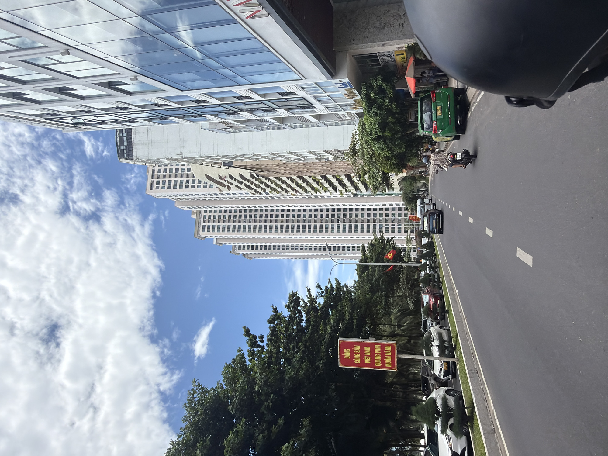 The image depicts a street scene in Nha Trang, Vietnam, characterized by tall buildings and lush trees lining the road. A red and yellow sign in Vietnamese, likely a political slogan, is visible on the left side of the road, next to a row of parked white cars and decorative trees. Further down the street, more cars and a green taxi are observed, with a person on a scooter navigating the traffic. The architecture features modern high-rise buildings with reflective glass facades, contrasting with the natural elements of the trees. The sky is partly cloudy, suggesting an outdoor urban environment on a bright day. The perspective suggests the image was taken from a moving vehicle, possibly a motorbike.