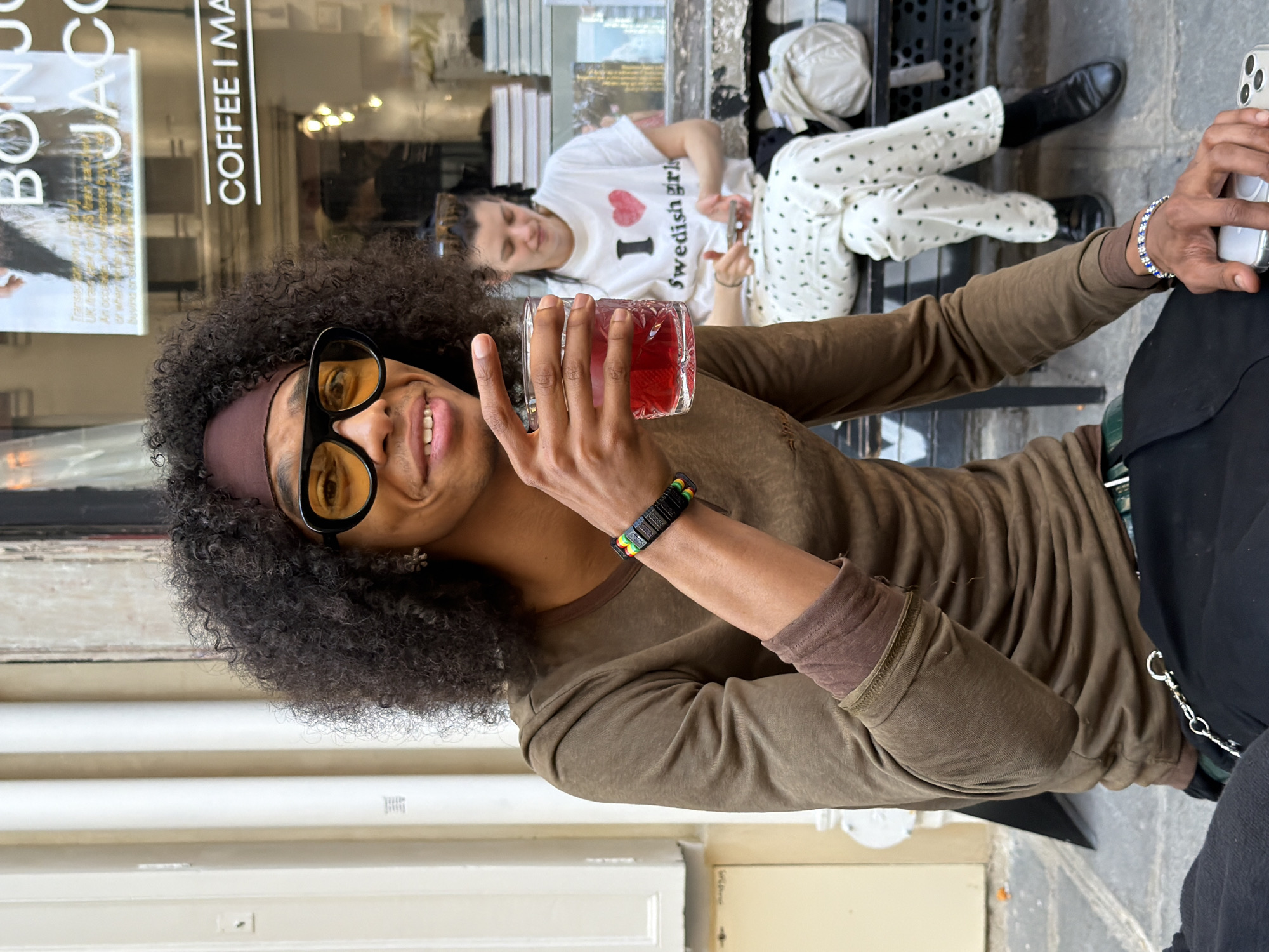 The image shows a man with curly hair and yellow-tinted glasses holding a glass of red liquid, likely a drink, in front of a cafe window. The cafe window has the words "COFFEE I MADE" written on it and an ad next to it. Behind the man, through the cafe window, is another woman wearing a white t-shirt with a red heart and the words "I ❤️ Swedish girls." She appears to be lying on a bench. Another person, whose lower body is visible, is seated on the bench next to her, wearing white pants with black polka dots and black boots. The overall setting suggests an outdoor cafe in Paris, France.