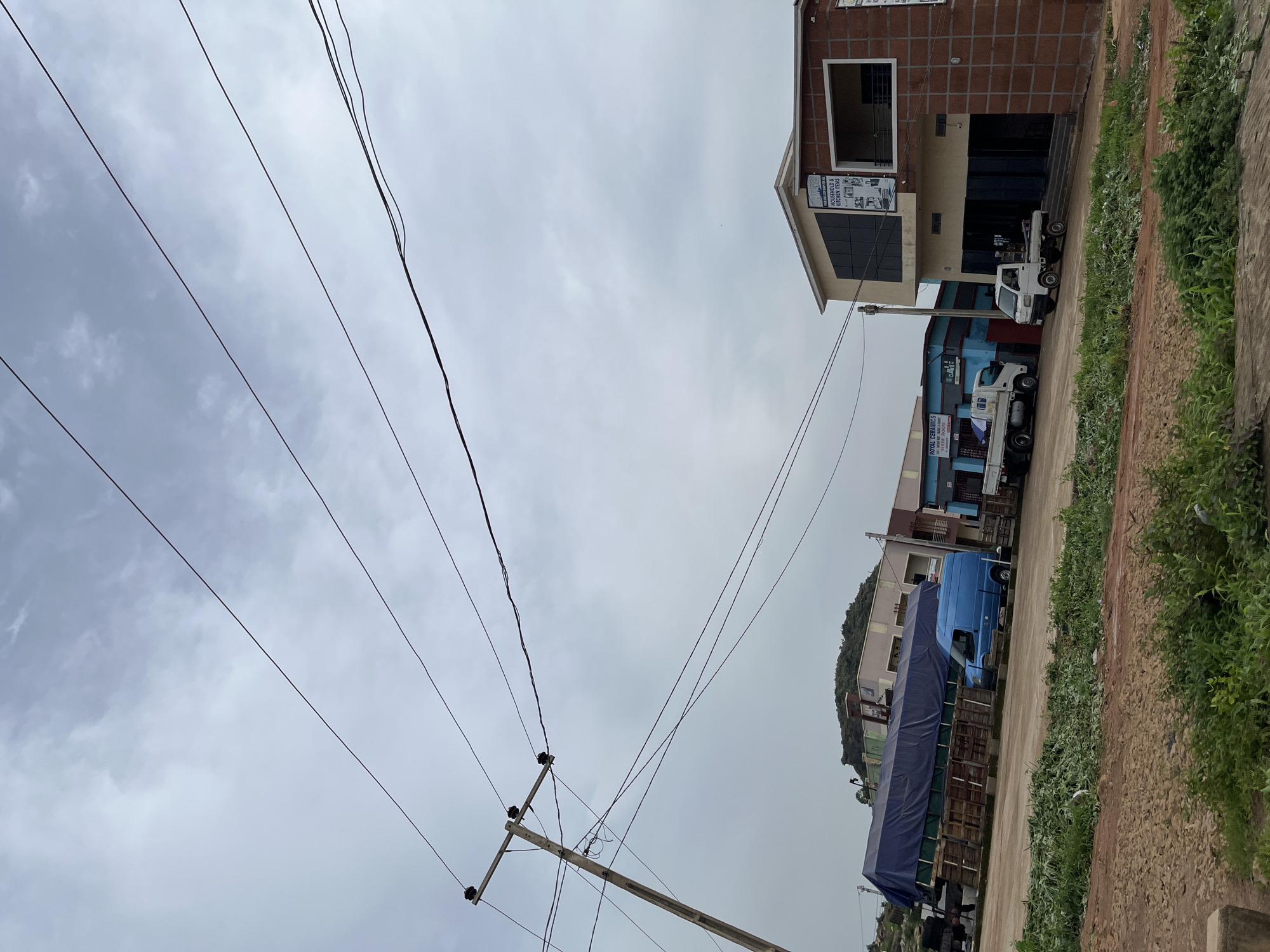 The image captures a street scene in Gura Top, Nigeria, under an overcast sky. A utility pole with multiple wires is prominently positioned on the left side of the frame. In the background, several buildings line the street, including a brick-faced structure with a modern glass facade, and other buildings with blue and beige exteriors. A blue cargo truck is parked in front of one of the buildings. In the foreground, a dirt road with patches of green vegetation runs along the bottom of the image. The overall scene depicts a commercial area with vehicles and buildings.