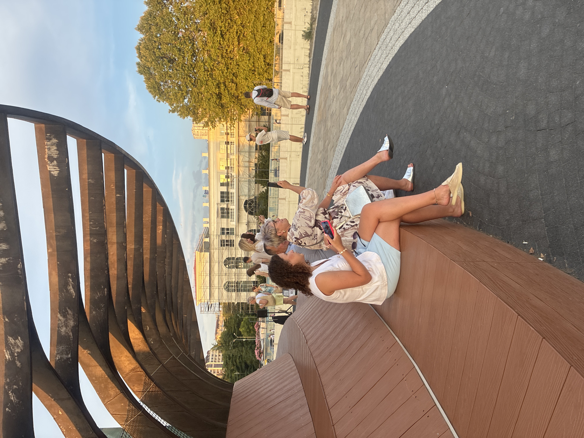 In Sochi, Russia, people are gathered on a wooden bench and a paved area beneath a modern, curved, metal canopy. In the foreground, two women are seated on the bench. The woman on the left wears a white sleeveless top and light blue shorts. The woman on the right is dressed in a floral-patterned dress and holds a smartphone. Behind them, several other individuals are visible, some standing and some walking. In the background, a building undergoing construction is situated next to a large, mature tree with golden foliage. The scene appears to be set during late afternoon or early evening, given the warm, golden light casting shadows.