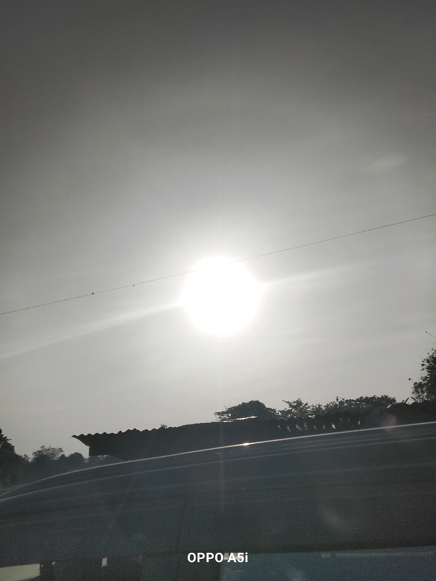 The image shows a bright sun in a hazy sky, likely during sunrise or sunset, casting a glare that makes it difficult to discern details. Silhouetted against the sky are the roofs of several structures, possibly houses or buildings, and a line of trees in the background. The foreground features the dark, reflective surface of a road or vehicle. A thin wire stretches across the sky with a small bird perched on it. The text "OPPO A5i" is visible in the bottom right corner, indicating the device used to capture the image. The scene is generic and does not provide specific visual cues to pinpoint its location within Tabanggele, Indonesia.