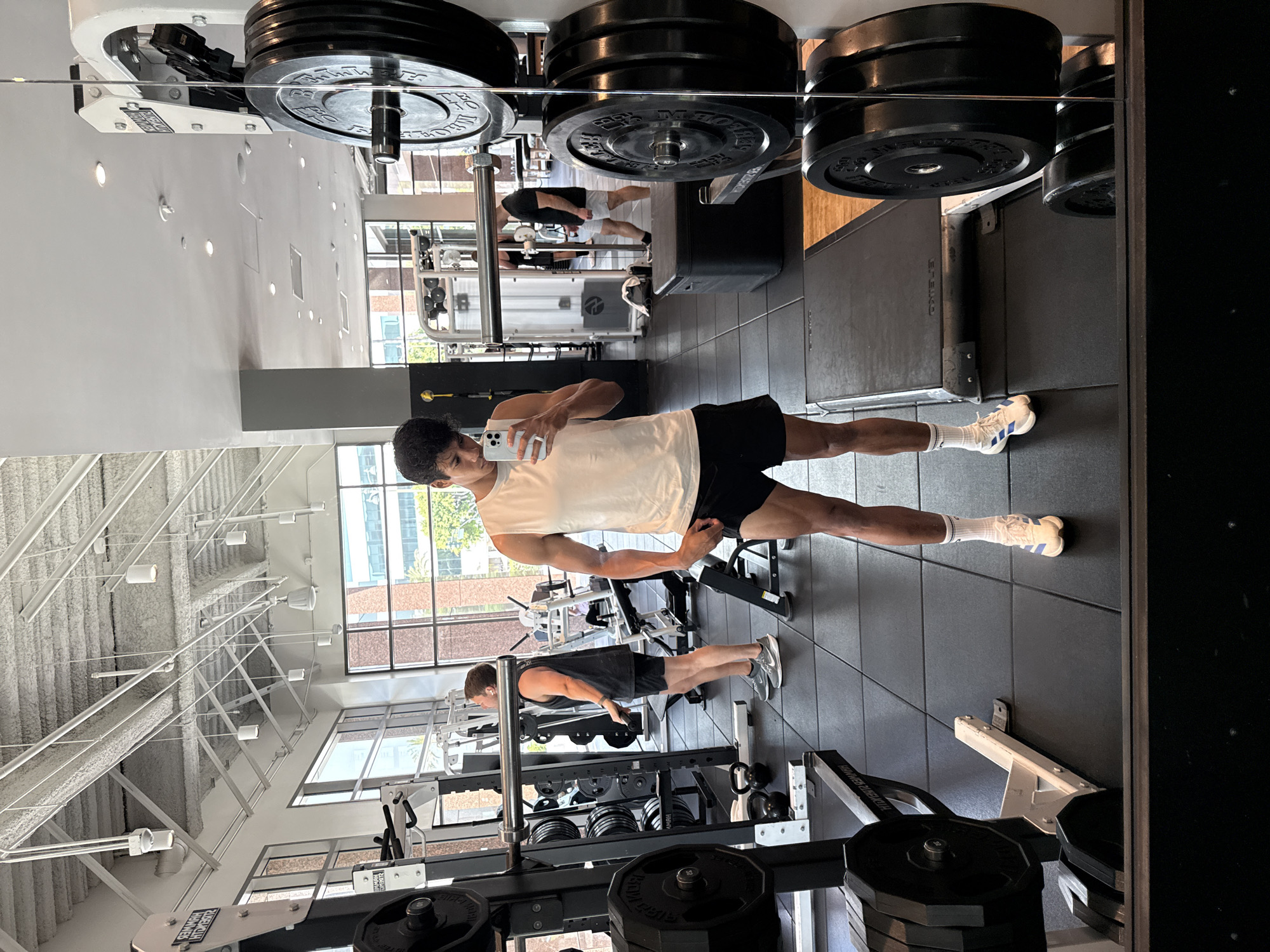 A young man takes a mirror selfie in a well-equipped, brightly lit gym. He stands centered in the frame, wearing a white sleeveless top, black shorts, white socks with blue stripes, and white athletic shoes, holding a white iPhone up to capture his reflection. His dark hair is neatly styled.
The gym features black rubber flooring and high white ceilings with exposed structural elements and modern recessed and track lighting. Large windows along the far wall allow natural light into the space, revealing parts of an urban environment outside, suggesting it is daytime.
In the background reflection, another man is visible, dressed in a black tank top and black shorts, engaged in an exercise near gym equipment. Various pieces of gym equipment are scattered around, including weight racks, barbells, benches, and machines. Text visible on equipment includes "Hammer Strength" and "ELEIKO," indicating specific brands. Some weight plates show numerical markings such as "25 KG" and "35 LB." The overall scene depicts a functional and modern fitness facility.