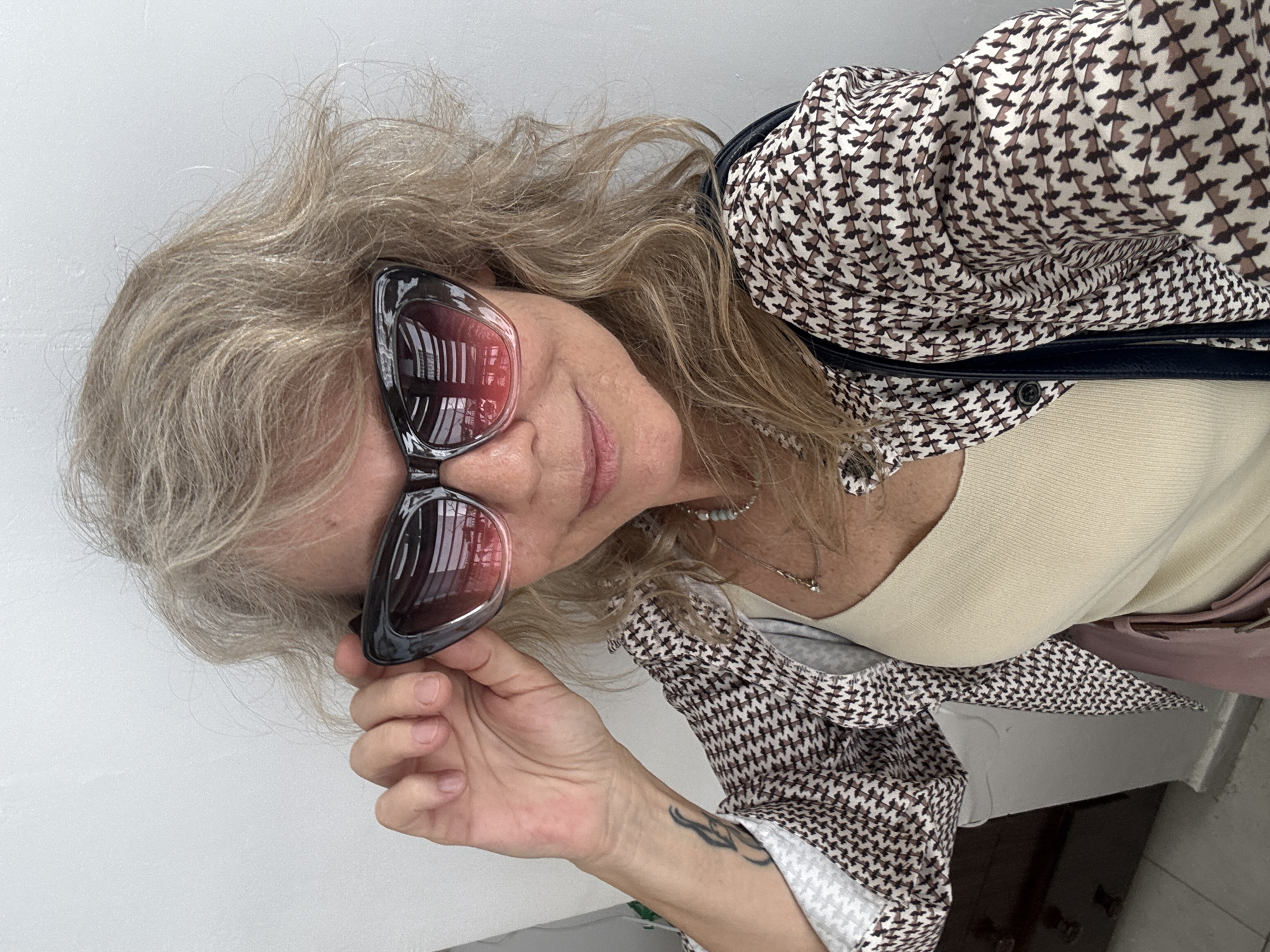 A middle-aged woman with blonde hair, wearing sunglasses and a patterned shirt, poses for a selfie against a white wall. Her left hand is raised, adjusting her sunglasses. A tattoo is visible on her left forearm. She wears a light-colored tank top underneath the shirt. The scene is indoors, with a hint of a dresser and tiled floor visible in the background. This image was taken in Ramat Gan, Israel.