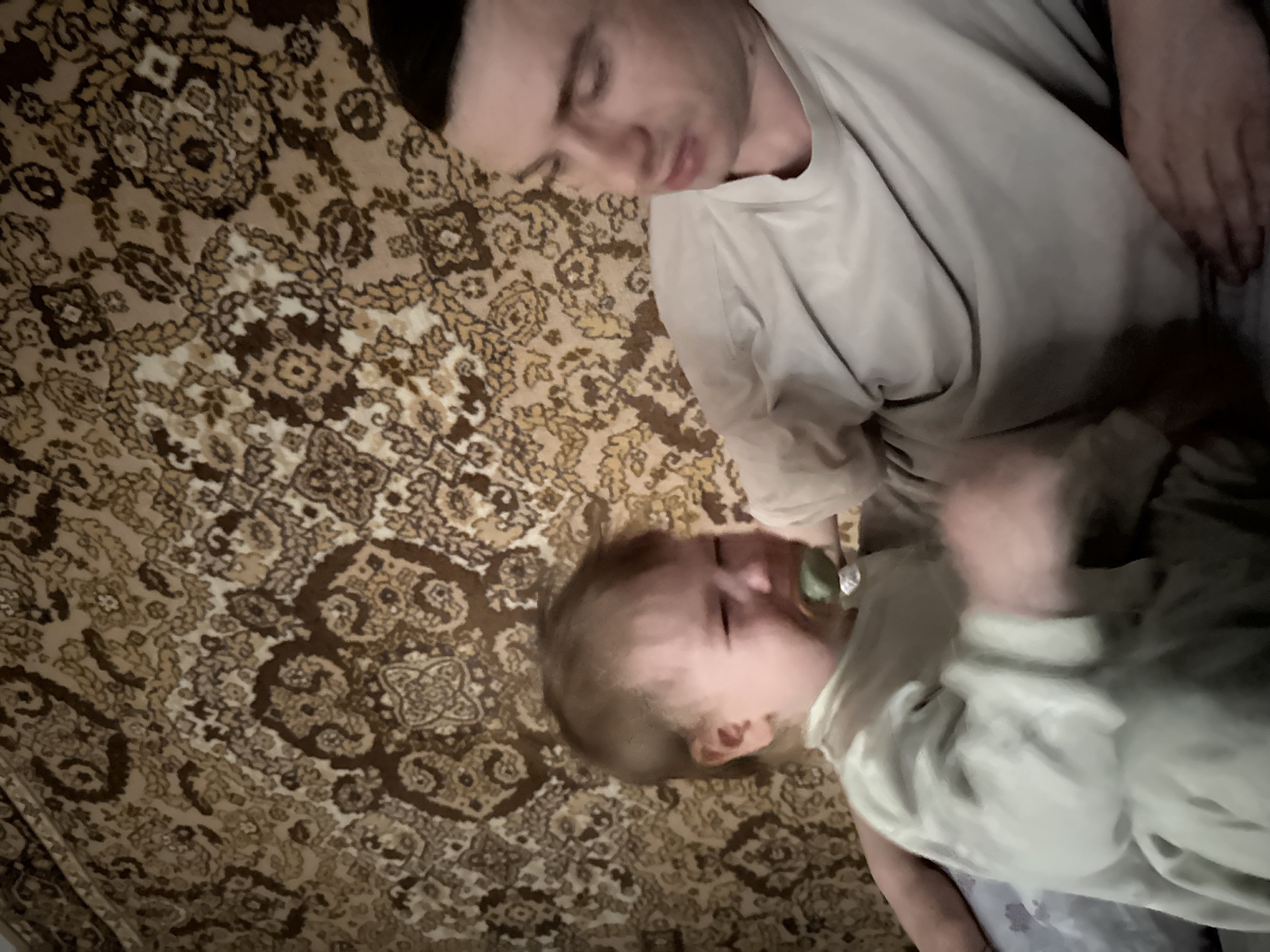 A man and a baby are lying on a patterned rug. The man, with short dark hair, is wearing a light-colored shirt. The baby has a pacifier in its mouth and is also wearing light-colored clothing. The rug has a complex, repeating floral pattern in shades of brown and beige. The image's location is unknown.