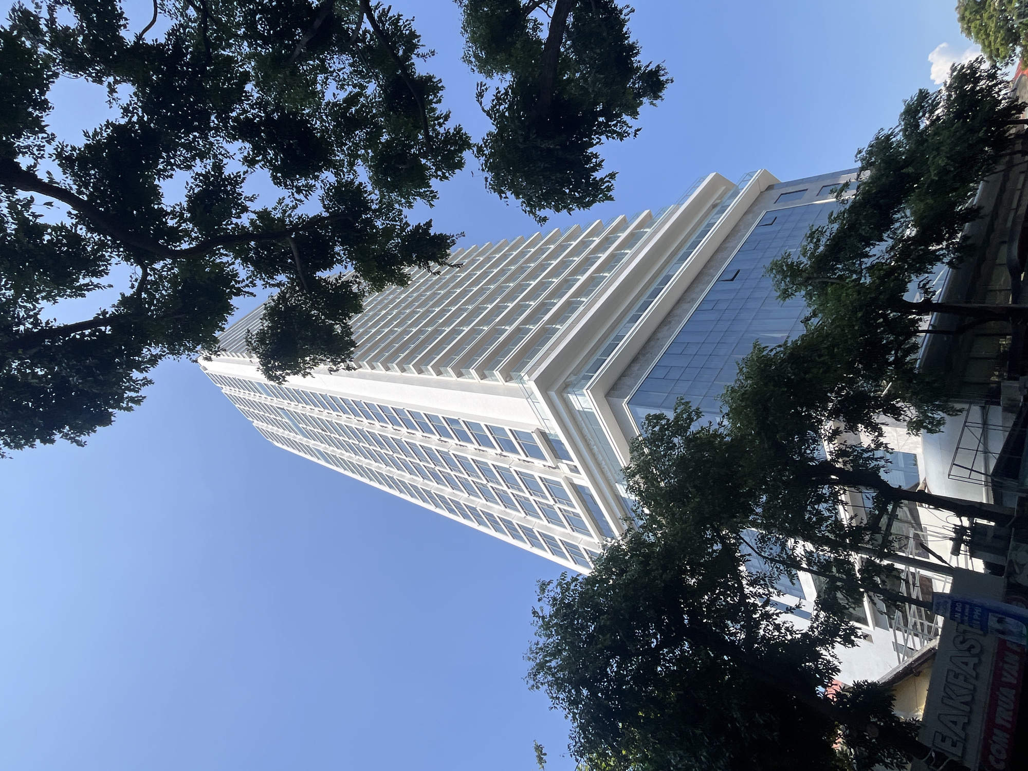 A low-angle shot captures a tall, white, modern building framed by trees against a clear blue sky. The building's facade features a grid of windows and balconies, with a section of glass panels on one side. The trees, with their dense green foliage, partially obscure the building, creating a foreground of shadows and leaves. In the lower right corner, a partially visible sign with text, including the word "BREAKFAST", hints at a street-level business. The location is Nha Trang Ward, Vietnam, identifiable by the architectural style and the signage. No people are visible.