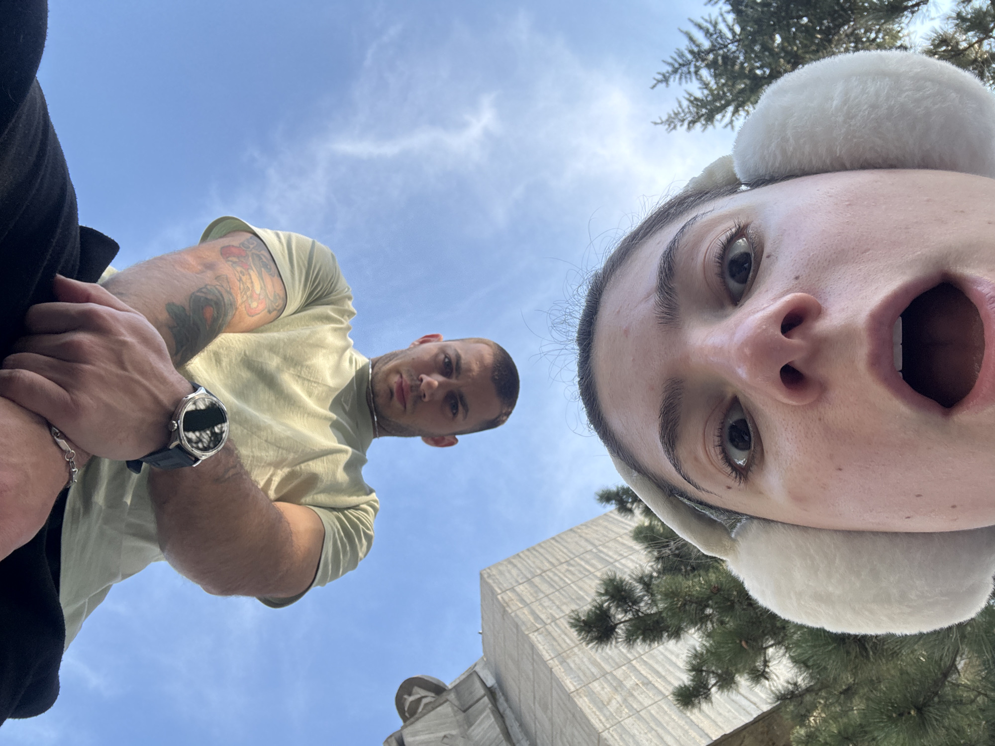 A worm's-eye-view photograph depicts two individuals in an urban setting with a clear blue sky. On the left, a man with a light green short-sleeved shirt, tattoos on his arm, and a watch on his wrist is visible. To the right, a woman wearing white earmuffs appears with her mouth open. A building and trees are visible in the background.