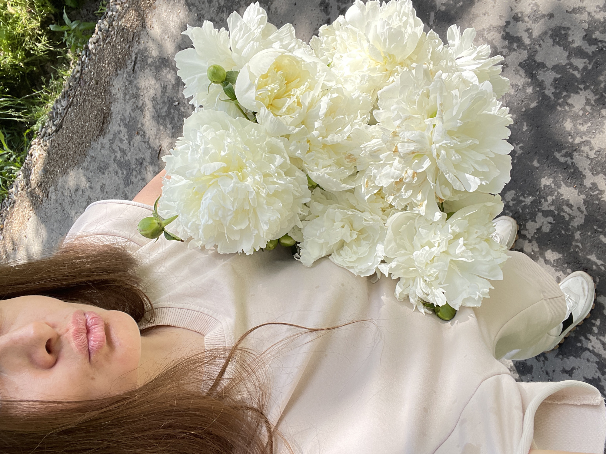 A woman with brown hair is holding a bouquet of white peonies in an outdoor setting. She is wearing a light-colored crewneck sweatshirt and light-colored pants, along with white sneakers with black accents. Her lips are pursed as if to give a kiss. The ground beneath her appears to be a paved surface with some gravel and vegetation to the right. The lighting suggests it is daytime. The specific location within the city of Заречный, Russia, is not identifiable from the visual information in the image.