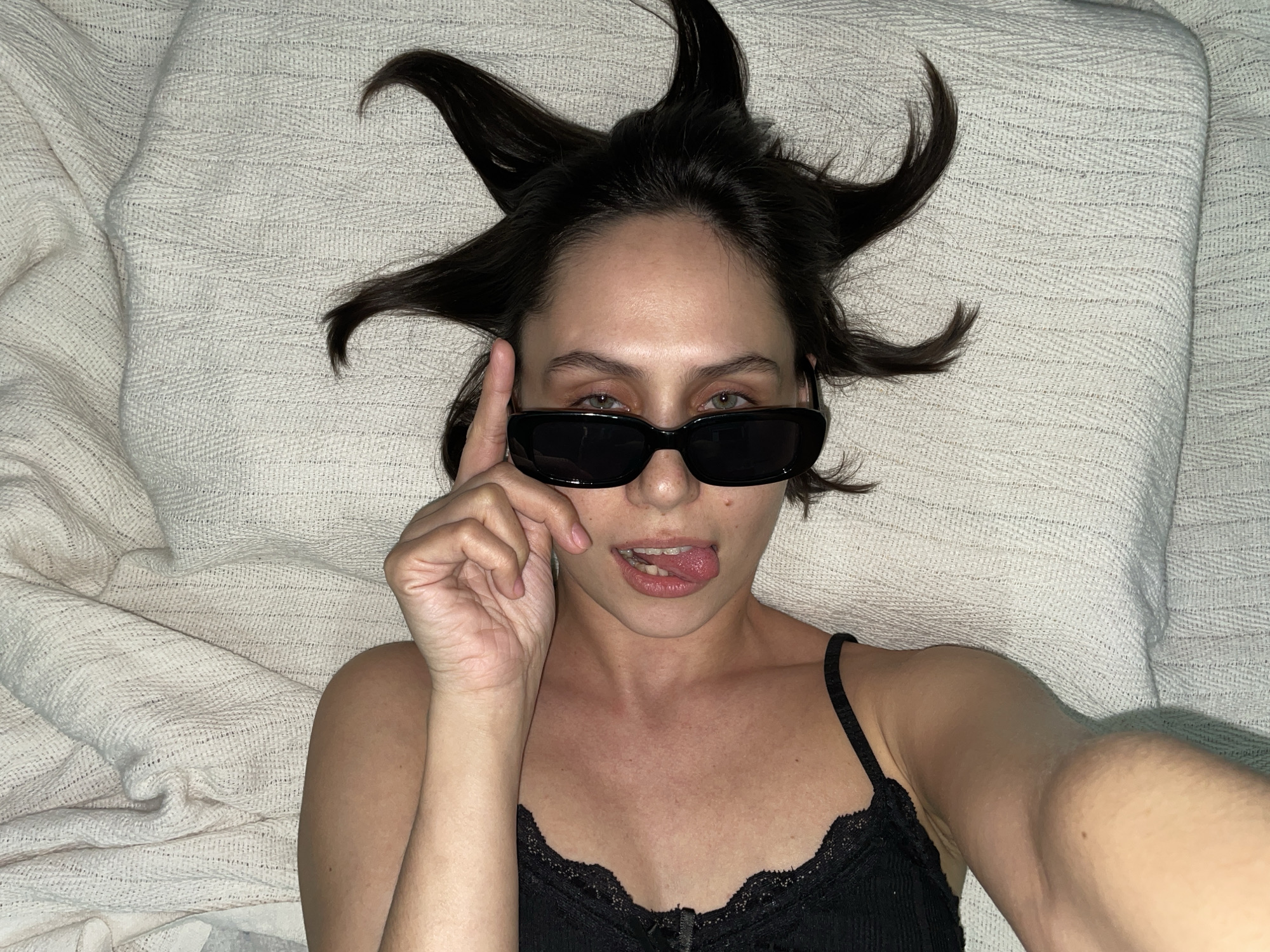 A young woman lies on a light-colored textured surface. She is wearing a black lace-trimmed camisole and black sunglasses. Her hair is styled to point outwards in several directions. She is holding up her right index finger near the sunglasses and sticking her tongue out. The environment appears to be indoors, possibly on a bed or sofa. No information indicates the location is Izhevsk, Russia.