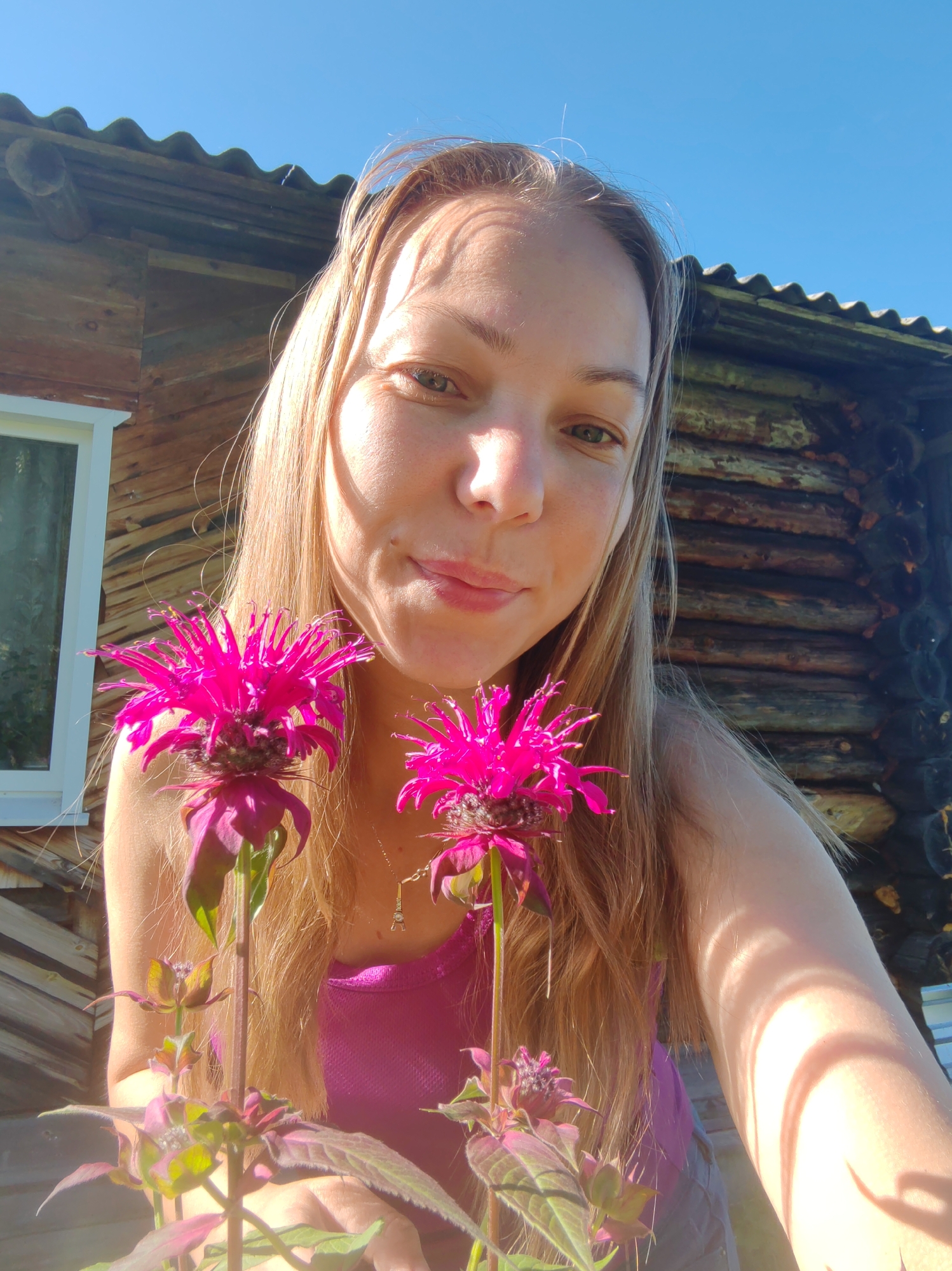The image depicts a young woman with long, blonde hair, holding two bright pink flowers in front of her face. She is wearing a pink tank top and grey shorts. The background shows a rustic wooden building, likely a house or shed, with a corrugated roof. The setting appears to be outdoors, with a clear blue sky visible. The woman is looking towards the camera with a slight smile. The overall scene suggests a casual, outdoor portrait taken on a sunny day. The location is specified as Chuchlem, Russia.