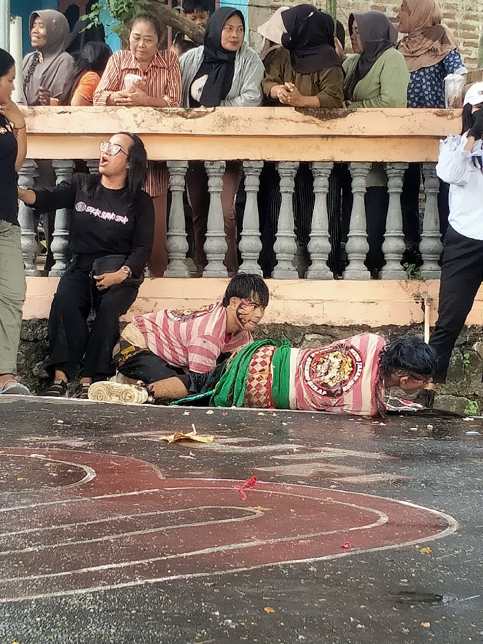 The image depicts an outdoor public display or performance in Jenggrik, Indonesia. The immediate foreground consists of a dark, wet ground surface, possibly asphalt, marked with painted red and white curvilinear lines and scattered with a yellow leaf-like object and small red debris.
Two individuals are on the wet ground. The person on the left is in a kneeling posture, facing right, wearing a pink and white striped short-sleeved shirt featuring a circular emblem on the back, dark pants, and a patterned fabric wrap around the lower body. Their face has dark markings, and dark streaks are visible on their face and dark hair. White athletic shoes are positioned near their left foot. The second individual, on the right, lies prone on their stomach, facing right, with their head lowered towards the ground. They wear a similar pink and white striped short-sleeved shirt with a circular emblem. Their torso and legs are partially covered and bound by a green fabric and a patterned textile. Their dark hair appears wet.
Above and behind the ground-level activity is an elevated concrete platform or balcony featuring a balustrade composed of light-colored concrete and grey decorative pillars. Approximately eight onlookers, primarily women, are positioned on this elevated platform, observing the scene below. Their attire includes various hijabs and casual clothing. One woman in an orange and white striped shirt holds a clear plastic cup. A child's head is partially visible among the spectators.
At ground level, to the left of the main subjects, a woman with dark hair and glasses, wearing a black long-sleeved shirt with white lettering (reading "SPAR SHARP SAP") and black pants, sits on the edge of the elevated platform's base. She is actively gesticulating with her right hand and holds a black bag on her lap. On the far right, partially visible at ground level, a person in a white long-sleeved shirt and a white cap stands observing the scene.