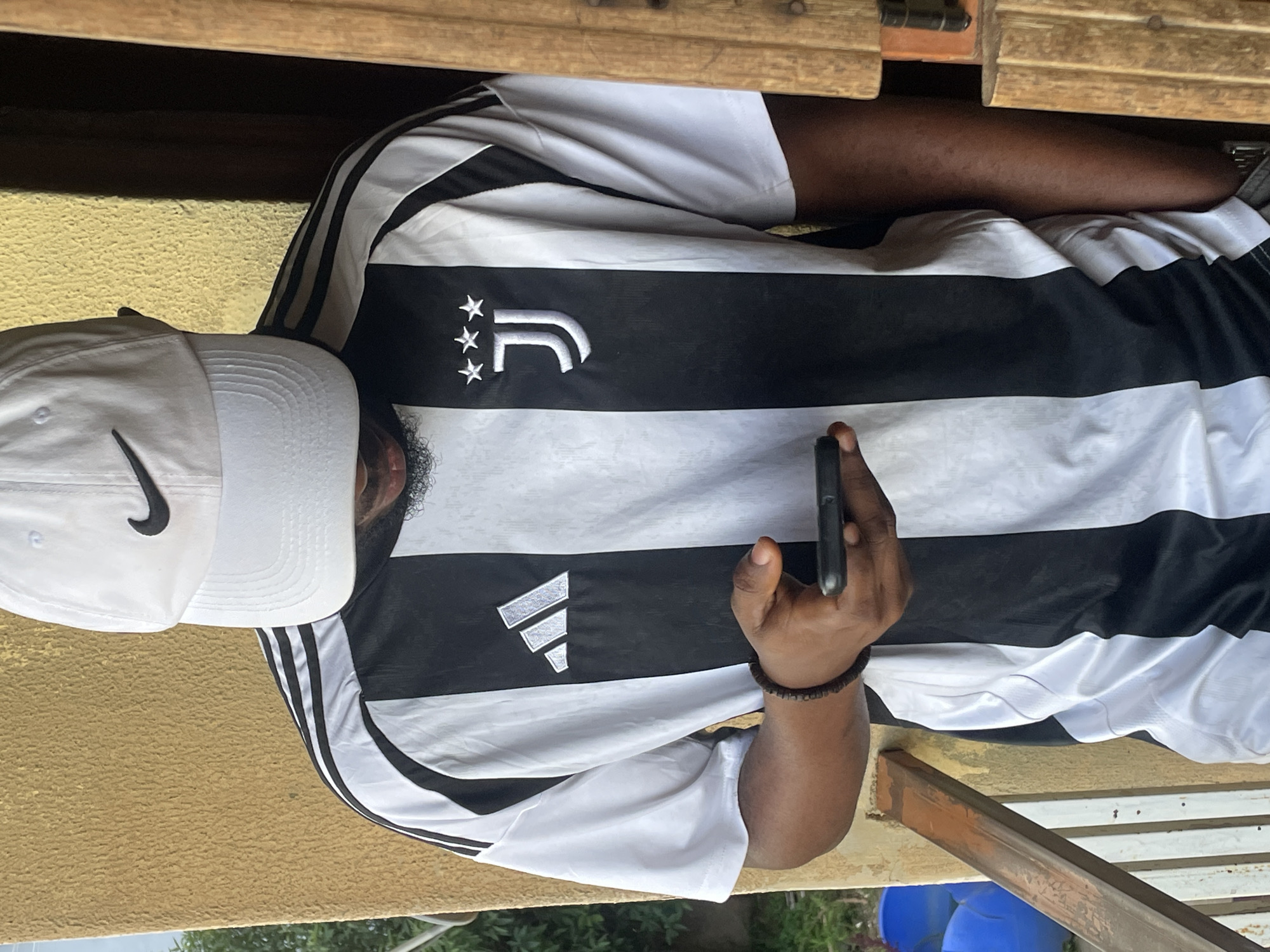 A person wearing a white Nike baseball cap and a black and white striped Juventus soccer jersey is holding a smartphone in their right hand, looking down at the screen. The person is standing outdoors next to a yellowish-brown textured wall, with a wooden beam to their right. The background shows some greenery and blue objects. The scene is likely in Jos, Nigeria.