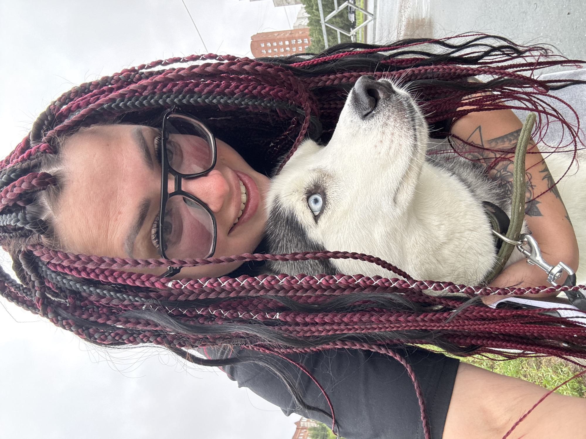 A selfie of a woman with dark, red-streaked braided hair and glasses holds a husky dog in the city of Tolyatti, Russia. The woman is wearing a black shirt, and the dog has striking blue eyes and white and black fur. They are outdoors with a blurred background of buildings and trees, suggesting a public space such as a park or street in Tolyatti. The overall impression is a casual, affectionate moment captured between the woman and her pet.