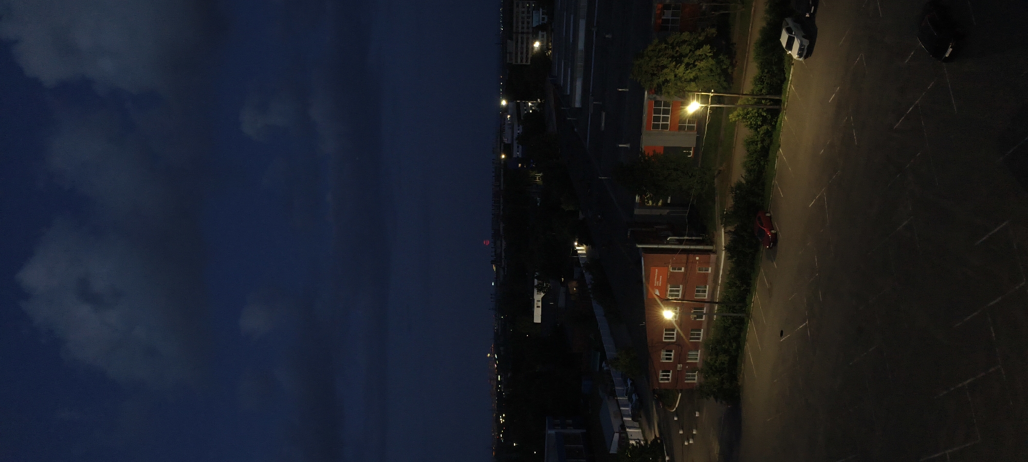 An aerial, vertical shot captures an industrial area at dusk. The sky is dark blue with heavy clouds, hinting at approaching night or stormy weather. In the distance, a city skyline is visible with scattered lights and a faint red glow. Closer to the foreground, several low-rise industrial buildings are depicted. One prominent red brick building has numerous windows and an illuminated sign. Adjacent to the buildings is a large, empty asphalt parking lot marked with white lines. A few cars are parked in the lot: a red sedan, a white SUV, and a dark-colored sedan. Streetlights illuminate sections of the parking lot and the surrounding area, casting long shadows. Trees and green bushes are interspersed throughout the scene, particularly along the edges of the buildings and the parking lot.