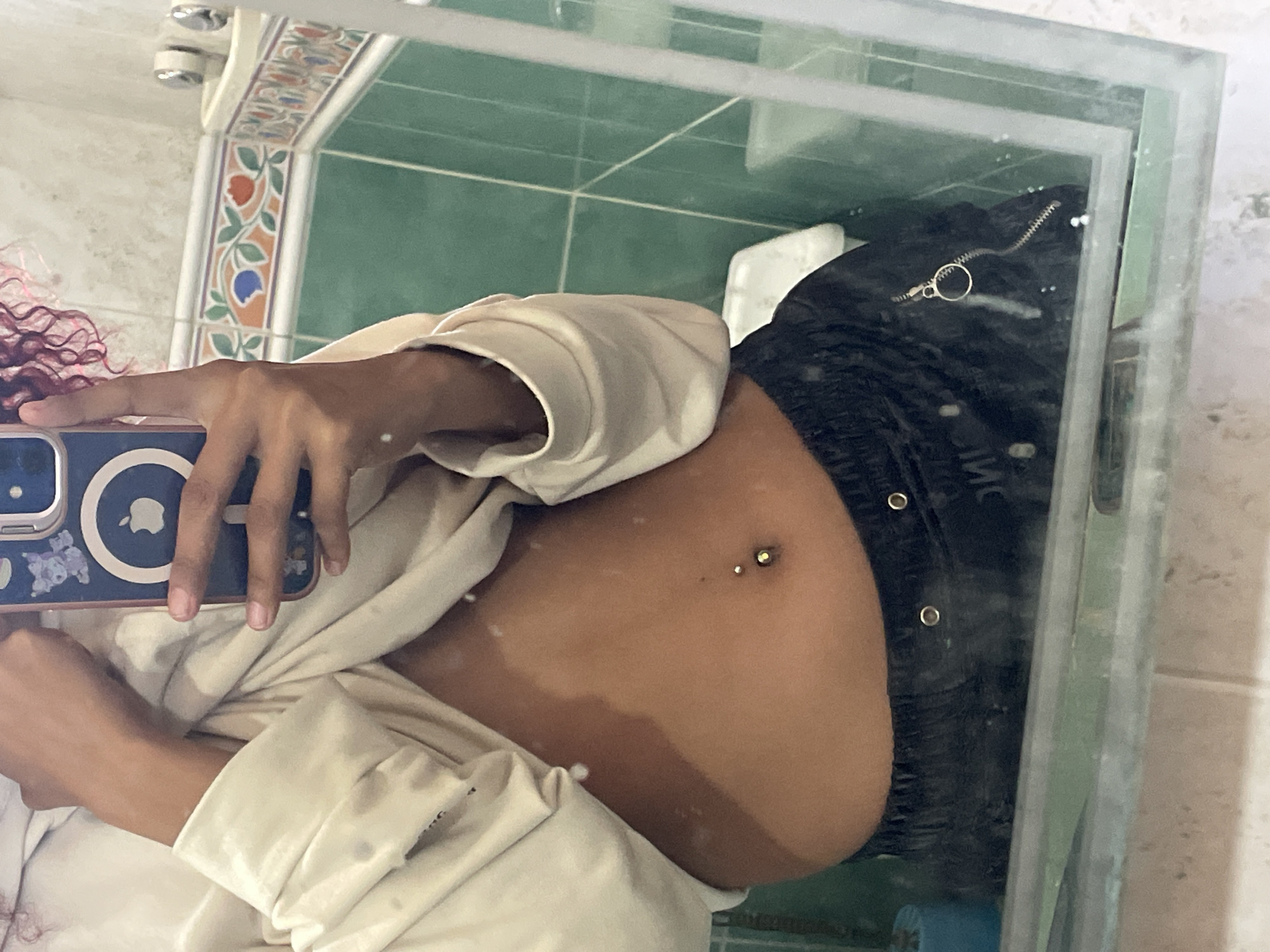 An indoor mirror selfie captures a person's midriff, hand, and smartphone, reflected in a bathroom setting. The person, likely female, has curly reddish-pink hair visible on the left. She holds a blue smartphone with a white Apple logo and small floral-like stickers on the case. Her right arm and hand are visible, and she wears a light-colored, long-sleeved top or jacket. Her abdomen is exposed, revealing a navel piercing with a double-ball barbell. She also wears dark-colored pants or shorts with a visible zipper and metal fasteners.
The background, seen through the reflection, consists of a bathroom interior. Green square tiles cover a wall, with a decorative border featuring white tiles adorned with floral patterns in red, blue, and green on an orange base. Chrome fixtures, possibly a shower head or faucet, are reflected at the top. A white ceramic edge, possibly part of a toilet or sink, is visible on the right. Layers of glass, suggesting shelves or shower partitions, are also reflected. The scene is located in Pedro González, Venezuela.