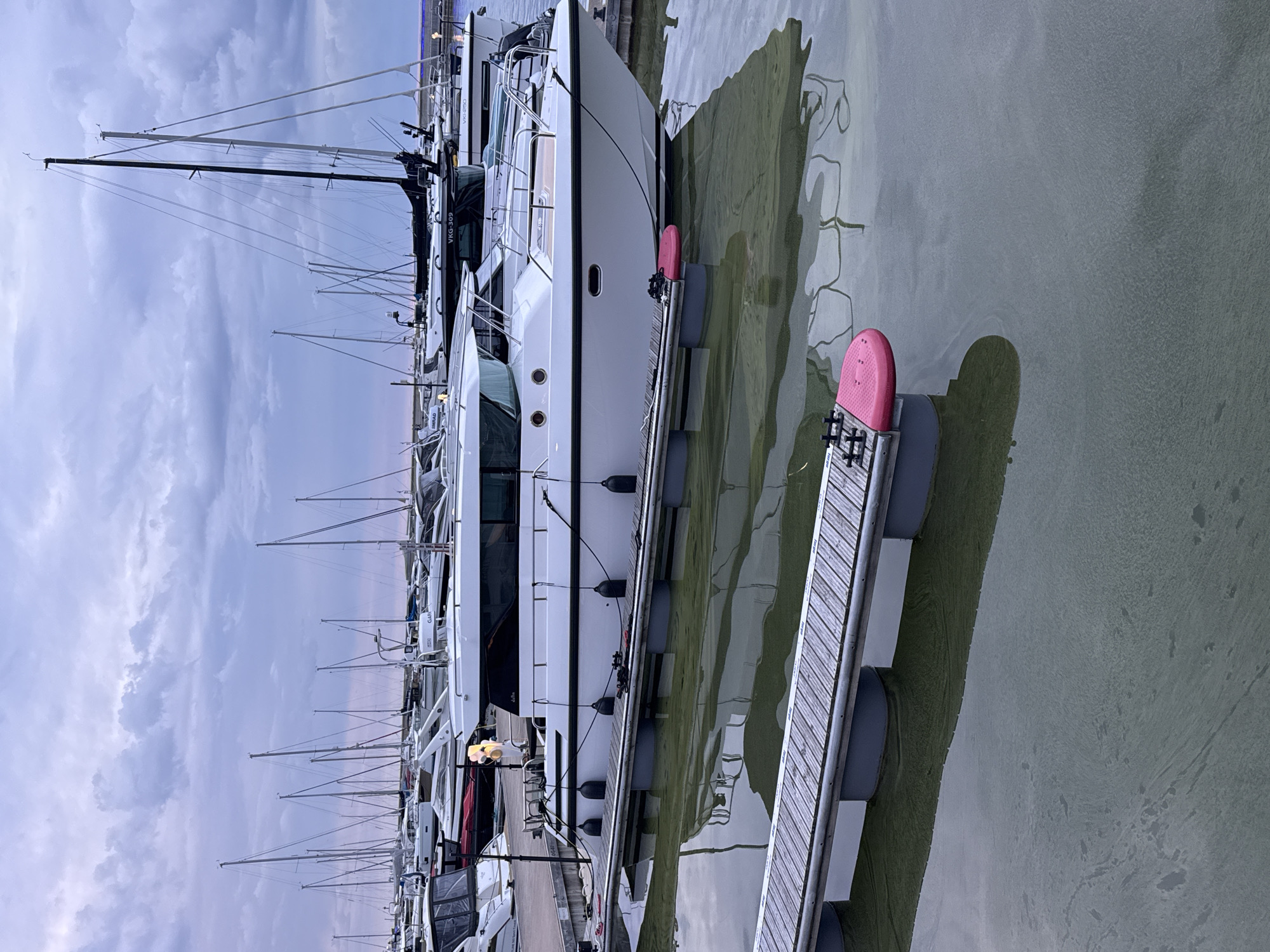In the city of Tallinn, Estonia, a marina hosts a fleet of docked sailboats and motorboats. The water, tinged with a greenish hue, reflects the overcast sky and the vessels. In the foreground, a wooden dock with gray flotation devices extends into the water, with a pink fender attached to its end. A white motorboat is moored to the dock, its hull detailed with black trim and windows. Several black fenders hang from its side. Behind this boat, other vessels are visible, including sailboats with tall masts, and more motorboats. The sky is filled with dramatic, dark clouds, suggesting approaching or recent rain. The overall scene is one of a quiet, overcast day at a harbor.