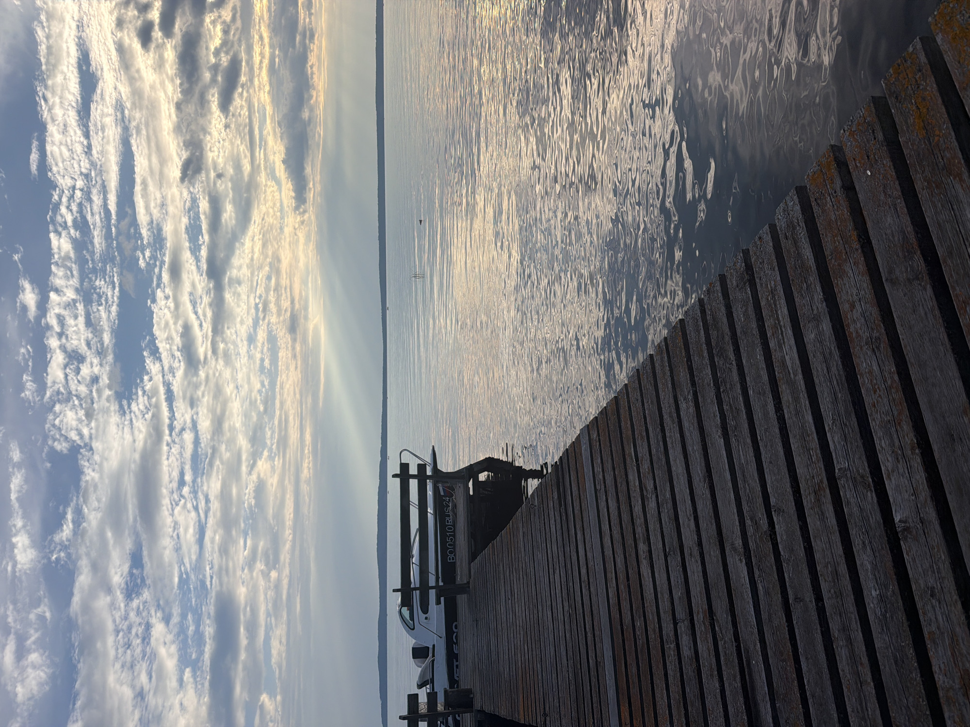 A wooden pier extends into a calm body of water under a cloudy sky. A white motorboat is docked at the end of the pier. The water reflects the light from the sky, creating shimmering patterns. In the distance, a tree line borders the horizon. The image appears to be taken in Stepanovo, Russia, based on the provided context.