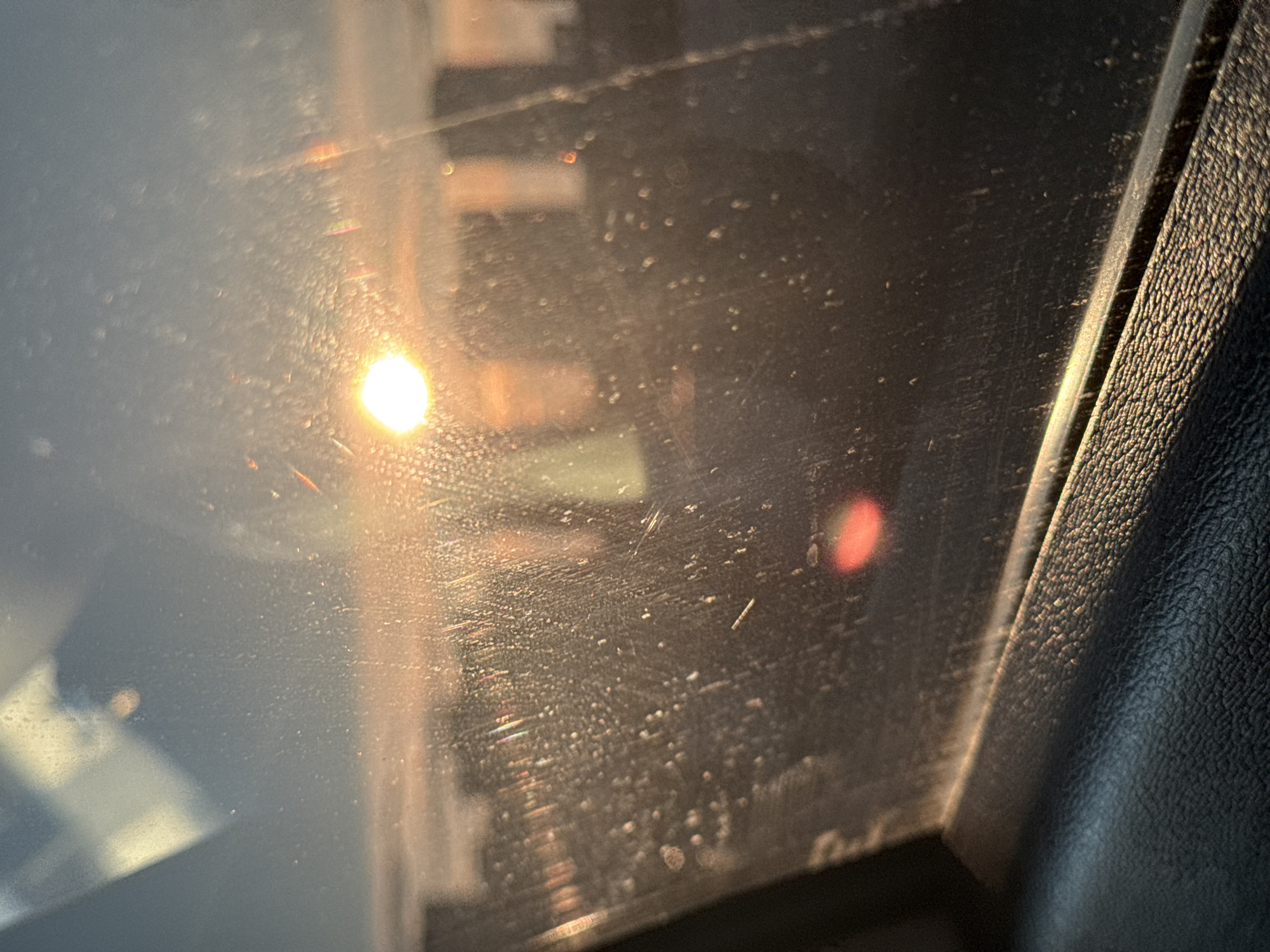 The image depicts a view through a dirty car window. Reflected sunlight obscures the outside scene, but there are hints of urban structures. Inside the car, a textured black surface is visible on the right, likely part of the dashboard or door. The window itself is smudged and streaked.