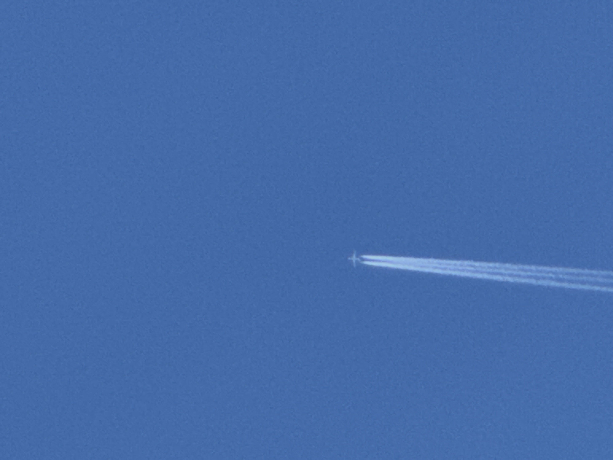The image shows a jet plane moving right across a clear, bright blue sky. The plane is leaving a long contrail behind it. There is no other visible detail beyond the jet and the sky. The location is unknown.