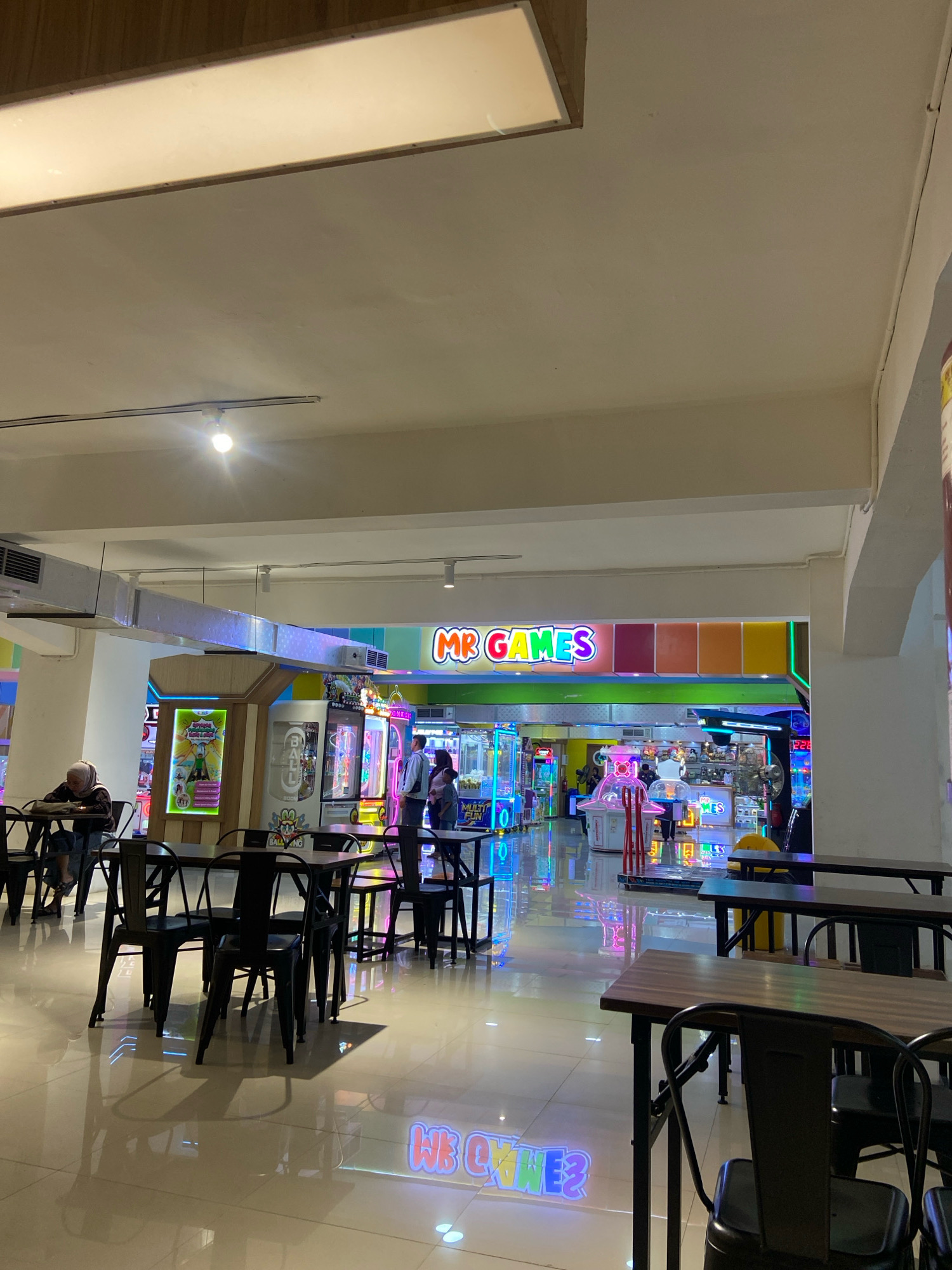 The image shows the interior of an arcade, identified by the prominent "MR GAMES" sign. The arcade features various game machines with colorful, illuminated displays, including crane games and other electronic amusement devices. The floor is polished and reflects the bright lights of the arcade. Several tables and chairs are arranged in a dining or waiting area in the foreground, suggesting that this is likely part of a larger entertainment complex or a mall in Kecamatan Pati, Indonesia. A few individuals are visible in the background, interacting with the games or walking through the arcade. One person is seated at a table in the foreground, while a small group stands near the game machines. The overall atmosphere is lively and illuminated by the arcade's neon lights.