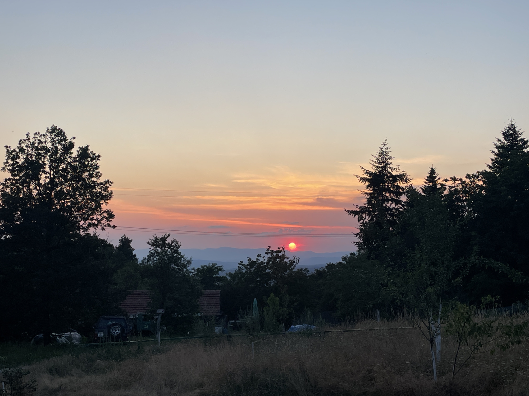 This is a photograph of a rural sunset scene, likely captured in or around Gabrovo, Bulgaria, given the architectural style of the distant structures and the natural landscape. The sun, a vibrant red orb, is low on the horizon, casting a warm glow across the sky. Wisps of clouds are illuminated in shades of orange and pink. Silhouetted against the colorful sky are the dark shapes of numerous trees, including several tall pine trees on the right side of the frame, and a broad leafy tree on the left.
In the middle ground, partially obscured by foliage, are a few simple buildings with red-tiled roofs, characteristic of rural Bulgarian architecture. A dark-colored SUV is parked in front of one of the buildings, suggesting human presence. The foreground is dominated by dry, tall grass and some low-lying vegetation, indicating a natural, undeveloped area. A thin fence runs across the middle ground, separating the immediate foreground from the area with buildings and vehicles. The overall atmosphere is tranquil and serene, typical of a rural evening at the end of the day. The photograph has been taken from an elevated position, looking down slightly on the scene.