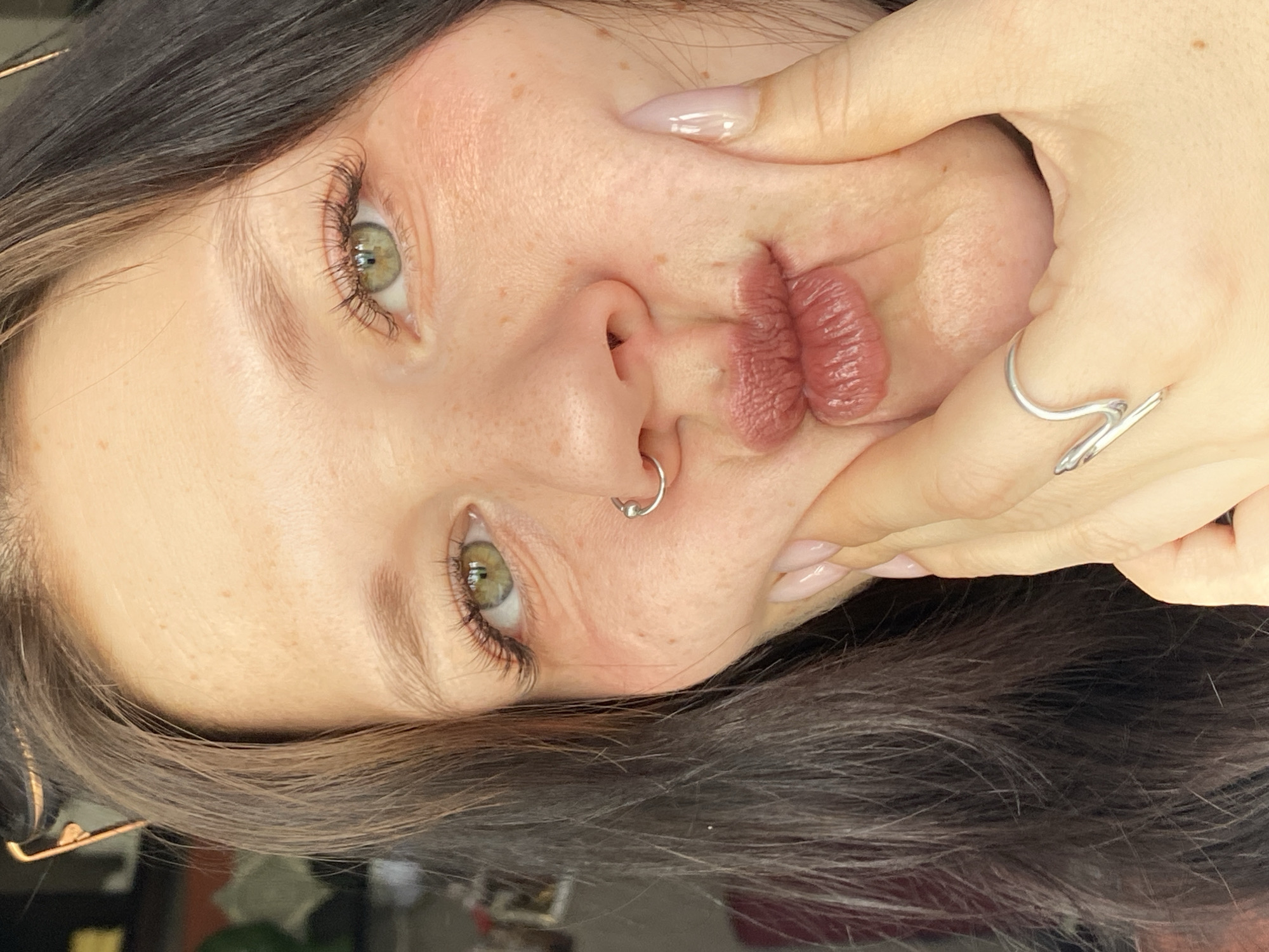 A close-up, portrait-style photograph shows a person with light skin, green eyes, and dark hair. The person has freckles on their face and a silver nose ring. Their lips are puckered and pressed against their nose, with a hand squeezing their cheeks. The hand has long, pale fingernails and wears a silver ring with a wave-like design. The background is blurry and out of focus. The location of the photograph is unknown, it cannot be determined from the provided image.