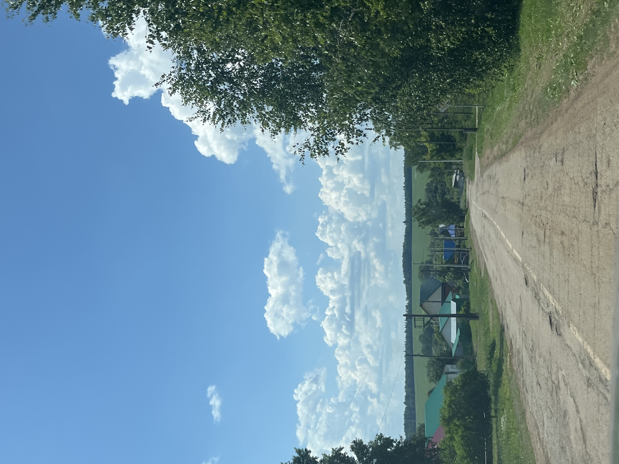 The image shows a view down a rural road in the village of Staryy Shagirt, Russia. The road is paved but appears to be in poor condition with visible potholes. On either side of the road, there are houses with green roofs, trees, and grassy areas. In the background, there are fields and a distant tree line under a bright blue sky with fluffy white clouds. The perspective suggests the photo was taken from a vehicle moving along the road. There are no people or animals visible in the image.