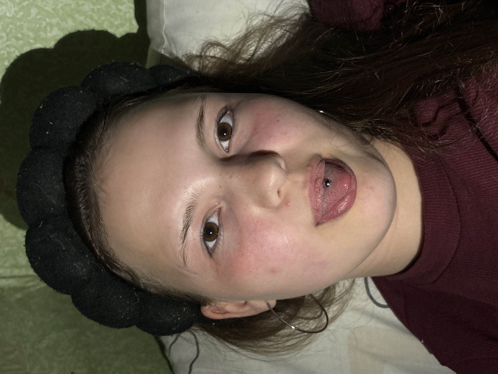 A young person with brown hair is lying on their back with a white pillow behind their head. They are wearing a black headband and a burgundy top. They have brown eyes and a tongue piercing. They are indoors. The background appears to be a light green surface. The city Фоки, Russia is not evident in the image.