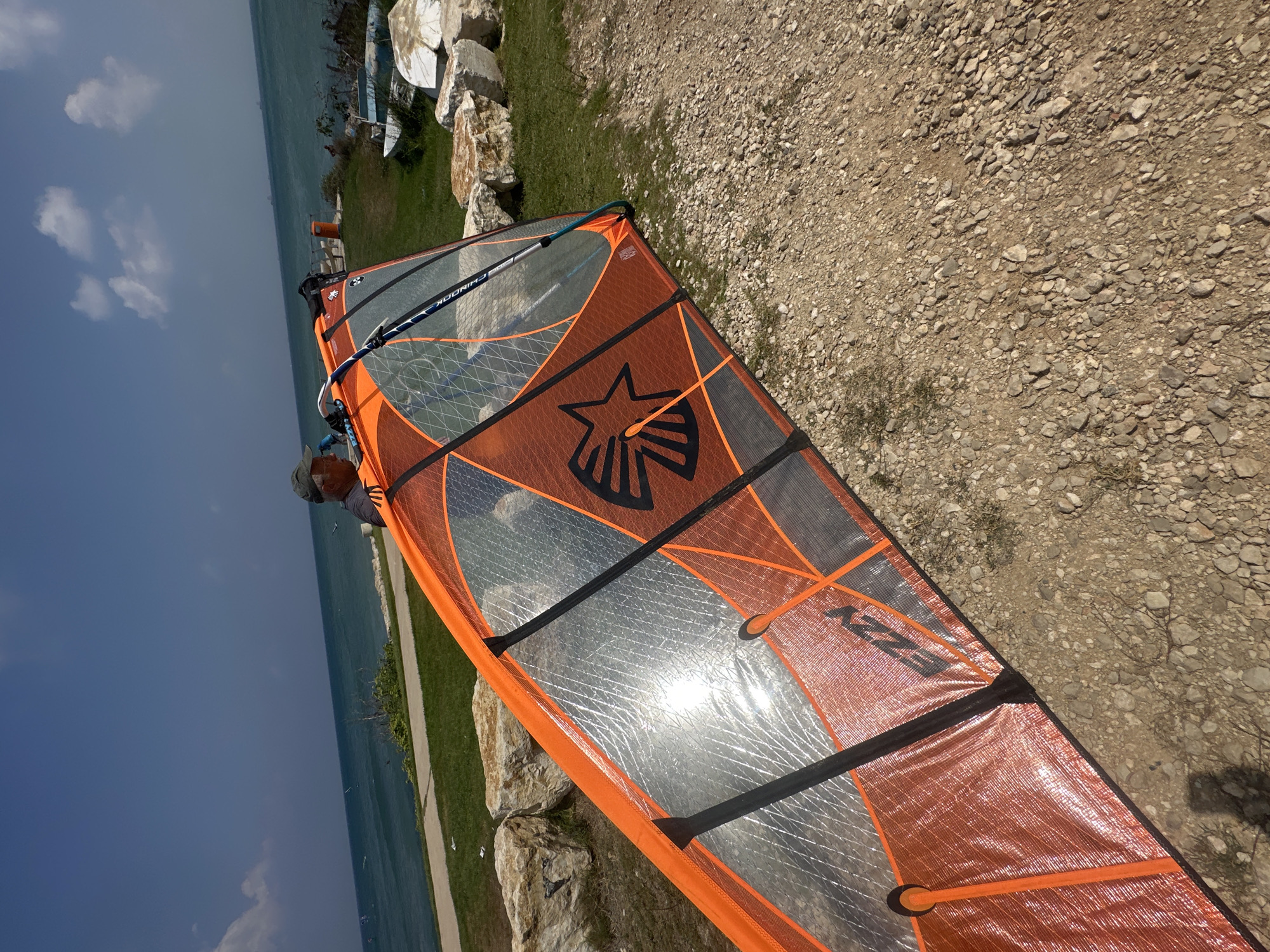 In Haifa, Israel, a windsurfing sail is lying on a gravelly area near a grassy patch and a rocky embankment. The sail is orange and translucent, with black and grey accents, and a black star logo. A silver boom is attached to the sail. In the background, a man wearing a cap is standing by the sail. Beyond the embankment, the blue sea stretches to the horizon under a bright, partly cloudy sky. There are a few boats visible on the water and some white boats pulled up on the shore. The sun is shining brightly, reflecting off the sail.
