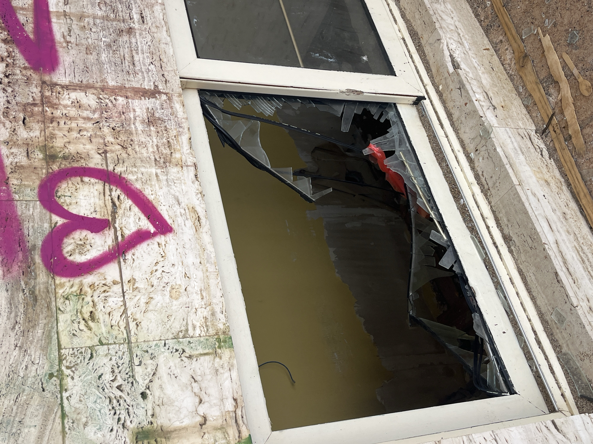 A broken window is visible in a building with graffiti on the exterior wall. The graffiti includes a heart shape sprayed in magenta. The window frame is white. The glass is shattered, with jagged edges and fragments remaining in the frame. The wall of the building is beige and has a rough texture. The location is Budva, Montenegro.