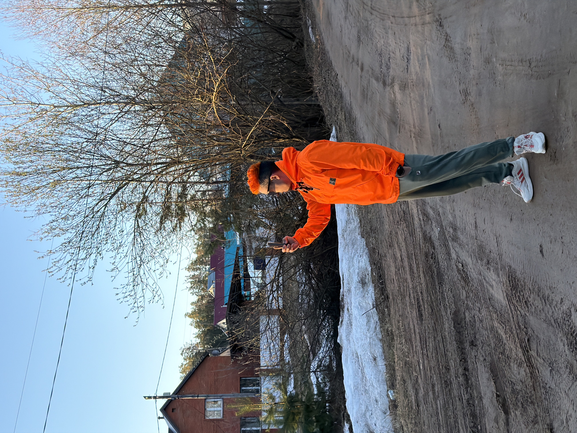 The image shows a man standing on a dirt road in what appears to be a rural or suburban area. The man is wearing an orange jacket, dark green pants, and white sneakers. He has brightly colored orange hair and is holding a cell phone in his outstretched hand. In the background are residential buildings, a snow bank, and trees without leaves. The sky is blue with no clouds. The location is Chaikovsky, Russia.