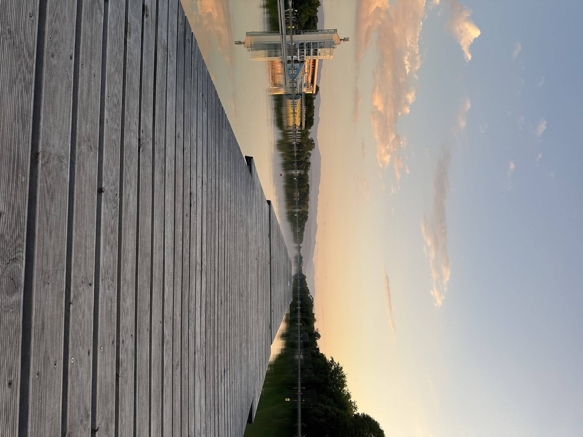 The image shows a dock extending into a body of water, likely a lake or river. The dock is constructed of wooden planks, with visible gaps between them. On the opposite side of the water is a building with a modern architectural design, partially reflected in the water. A line of trees is visible between the building and some mountains in the distance. The sky features clouds with a mixture of colors, including pink, orange, and blue, indicating either sunrise or sunset. The image is likely taken in Plovdiv, Bulgaria, given the building's reflection on the water is the Plovdiv Rowing Channel's main building.