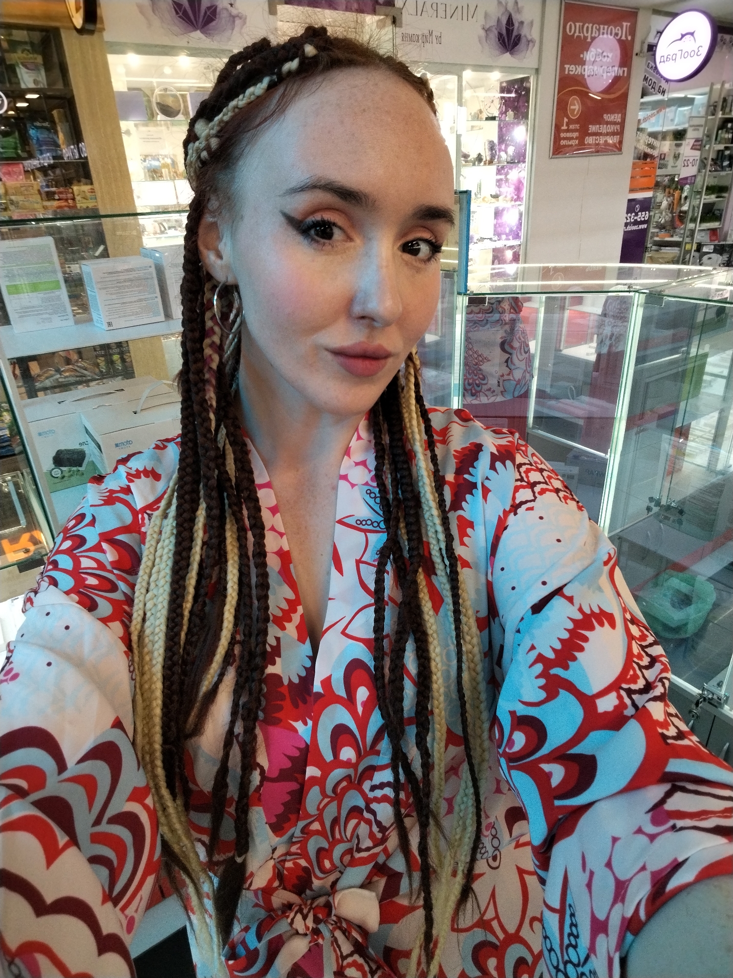 The image shows a woman with a fair complexion and freckles, wearing brown and blonde braids, posing for a selfie indoors. She has distinct eyeliner and hoop earrings. She is wearing a colorful robe with a floral pattern in shades of red, white, and blue. The background appears to be a store, indicated by the glass display cases and merchandise shelves. A sign is visible with some of the text readable in reverse and/or in Russian characters, including the words "МОД БН" and "LNUSOIN DKGL". Based on the text "moto" on one of the boxes visible through the glass, and the sign indicating "sioes.www", it's possible that this is a electronics or mobile phone store. A sign with a stylized image of a rabbit is also visible, in reverse. Another sign has readable text on it: "022-353 10-55." The location is Izhevsk, Russia.