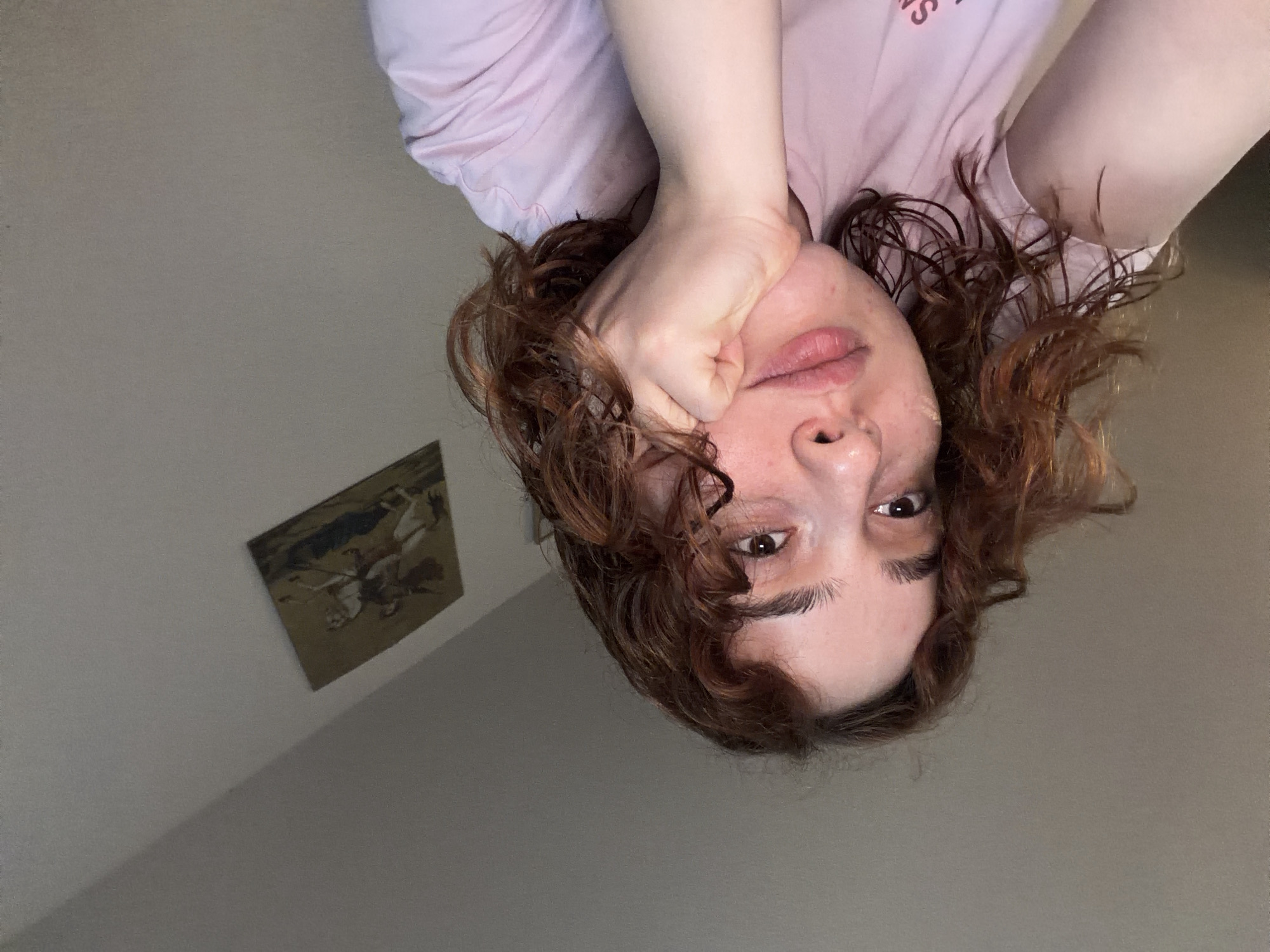 The image is a close-up, slightly blurry, selfie of a young woman with curly auburn hair and a pensive expression. She is resting her chin on her hand, wearing a light pink t-shirt. In the background, there is a wall with a framed tapestry depicting figures on horseback. There is no indication of Strasbourg, France in the image.