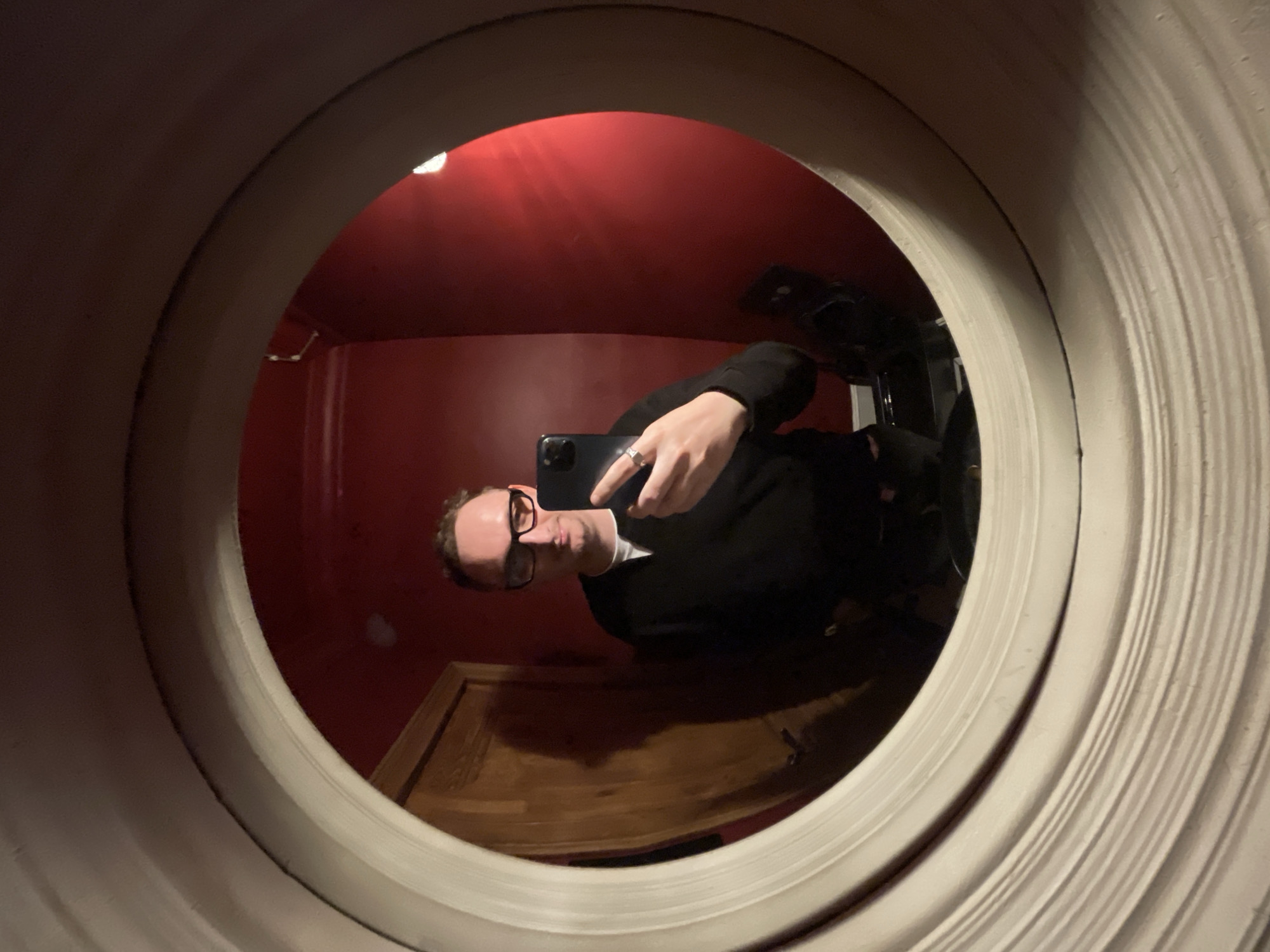 This is a fisheye mirror selfie taken indoors. The subject is a person with fair skin, short brown hair, and glasses, wearing a black jacket over a white shirt. They are holding a smartphone to take the picture. The person is making a peace sign with their left hand. The mirror is framed by a textured, light-colored border. The background reflected in the mirror shows a room with red walls, a wooden surface that appears to be a desk or dresser, and a dark object, possibly a chair or cabinet. There are also reflections of light on the wall, suggesting overhead lighting. The fisheye lens distorts the image, making the edges curve outwards. Based on the clothing and indoor setting, it is likely not daytime and the weather is irrelevant. The scene appears to be a personal space, possibly a bedroom or home office. There is no visible text in the image.