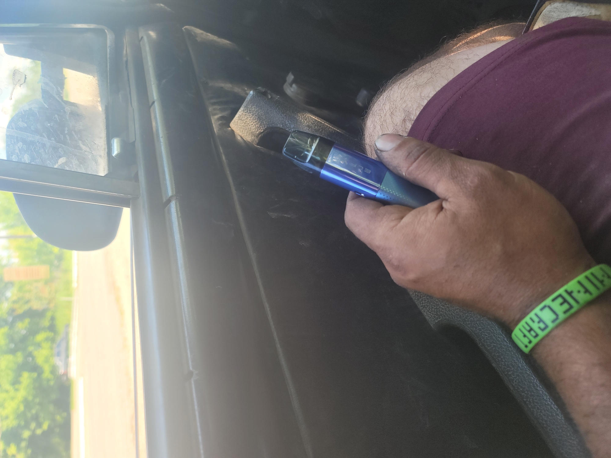 A person's hand, holding a blue vape pen, is shown from the wrist up. The hand is resting on a person's leg, covered by a maroon fabric garment. A green wristband with the word "MINECRAFT" in white letters is visible on the person's wrist. The background appears to be the interior of a vehicle, with a window showing a blurred outdoor scene. The image does not provide information about the location in Pereshchepyne, Ukraine, or any specific details of the surrounding city.