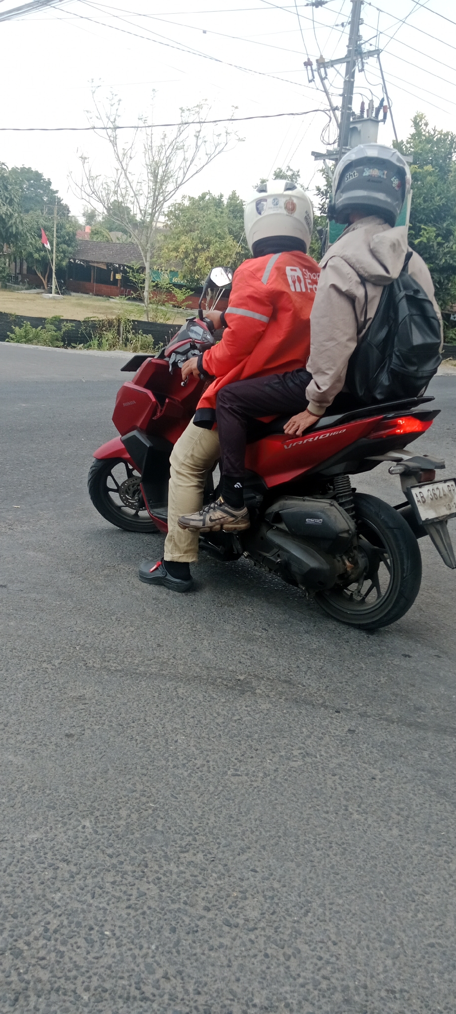The image shows two individuals riding on a red scooter in Maguwoharjo, Indonesia. The person in the front is wearing a red jacket with "Shopee Food" logos and a white helmet. The person in the back is wearing a light brown hooded jacket and a grey helmet. The scooter has a license plate that reads "AB 3624 PP". In the background, there are trees, a building with a thatched roof, and power lines. The scene appears to be on a paved road.