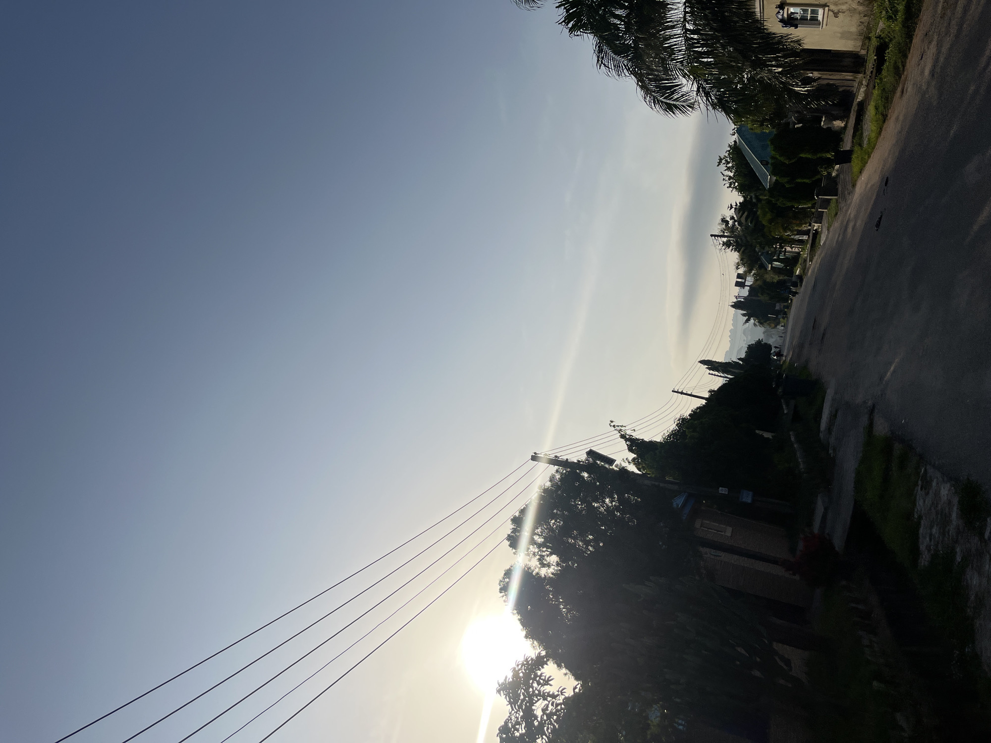 This image depicts a residential street in Abuja, Nigeria, during what appears to be early morning or late afternoon, judging by the low angle of the sun. The sky is a pale blue, transitioning to a lighter hue near the horizon. Several power lines are strung across the upper left portion of the frame, casting shadows.
The street itself is paved and slopes gently uphill into the distance. On the left side of the street, there are trees and foliage, with some residential buildings partially visible behind them. These buildings appear to have brickwork. On the right side of the street, more trees, including a prominent palm tree, line the road. Residential buildings with windows and roofs are visible, with one house in the foreground displaying a distinct window with white trim. The environment is lush with greenery, suggesting a well-maintained residential area. The overall impression is one of a quiet, suburban street scene bathed in soft, natural light.
