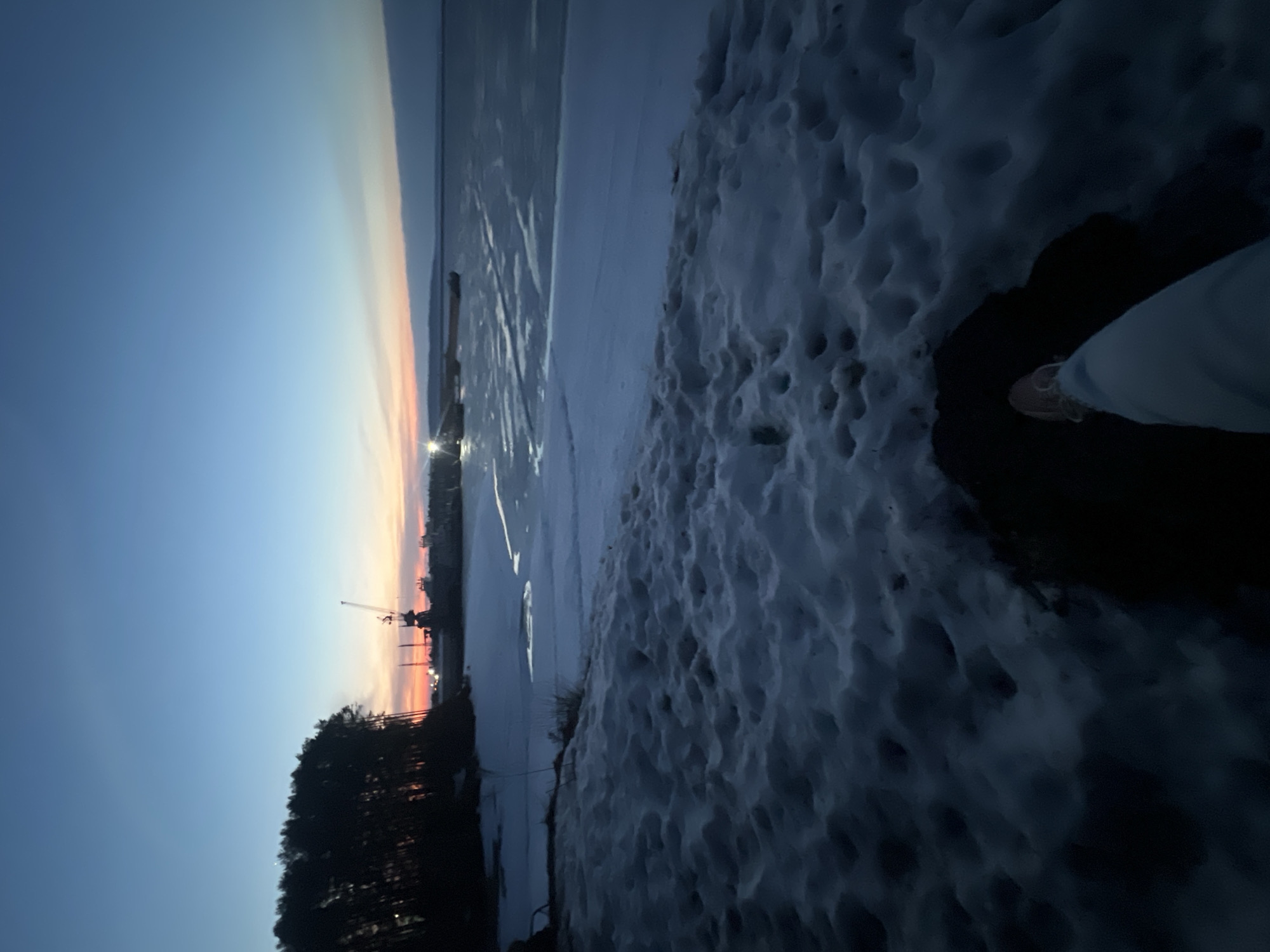 The image depicts a winter scene in Chaikovsky, Russia at dusk. The sky transitions from blue to orange near the horizon, indicating a sunset or sunrise. A frozen body of water, likely the Kama River, stretches towards the horizon. Along the shore is a snow-covered area with foot prints. The silhouette of a coniferous forest borders the shoreline to the left and a crane is silhouetted against the sunset. The picture includes an edge of a shoe and pant leg.