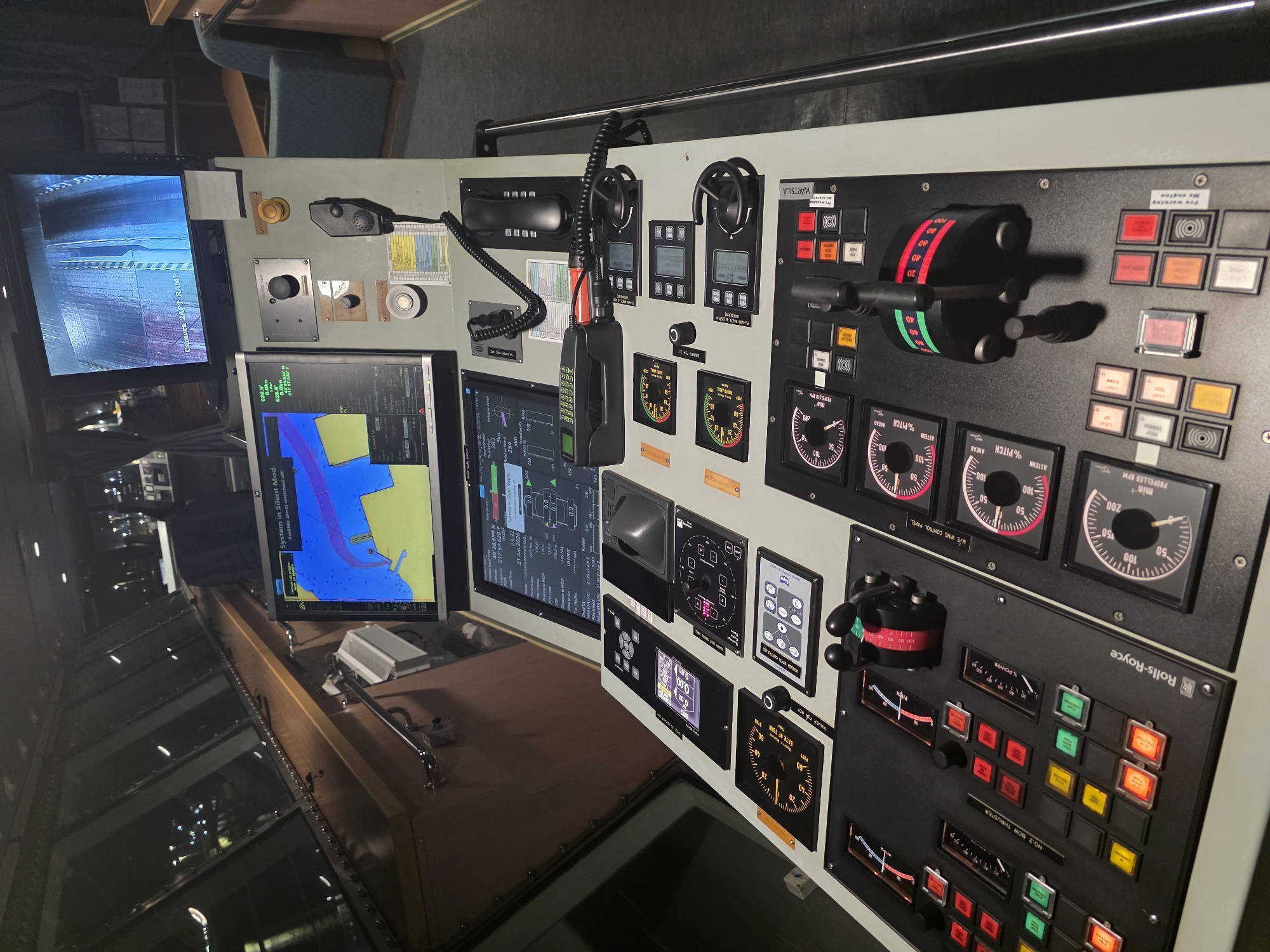 The image shows the bridge of a ship, likely a ferry or a commercial vessel, at night. The bridge is equipped with multiple screens displaying navigational data, charts, and camera feeds. There are numerous control panels with buttons, levers, dials, and gauges for operating the ship's machinery and systems, including thrusters and propulsion. A ship's telegraph is visible, indicating speed and direction commands. A telephone handset and a microphone are also present. Outside the windows, lights of a port or city can be seen, suggesting the ship is docked or near a port. The presence of the "Rolls-Royce" logo on one of the control panels indicates the use of their marine propulsion systems. The overall environment is functional and professional, typical of a ship's control center. The scene is dimly lit, with the primary illumination coming from the screens and control panel lights. There are no people visible in the image. The location is identified as Brindisi, Italy, suggesting the ship is in or near the port of Brindisi.