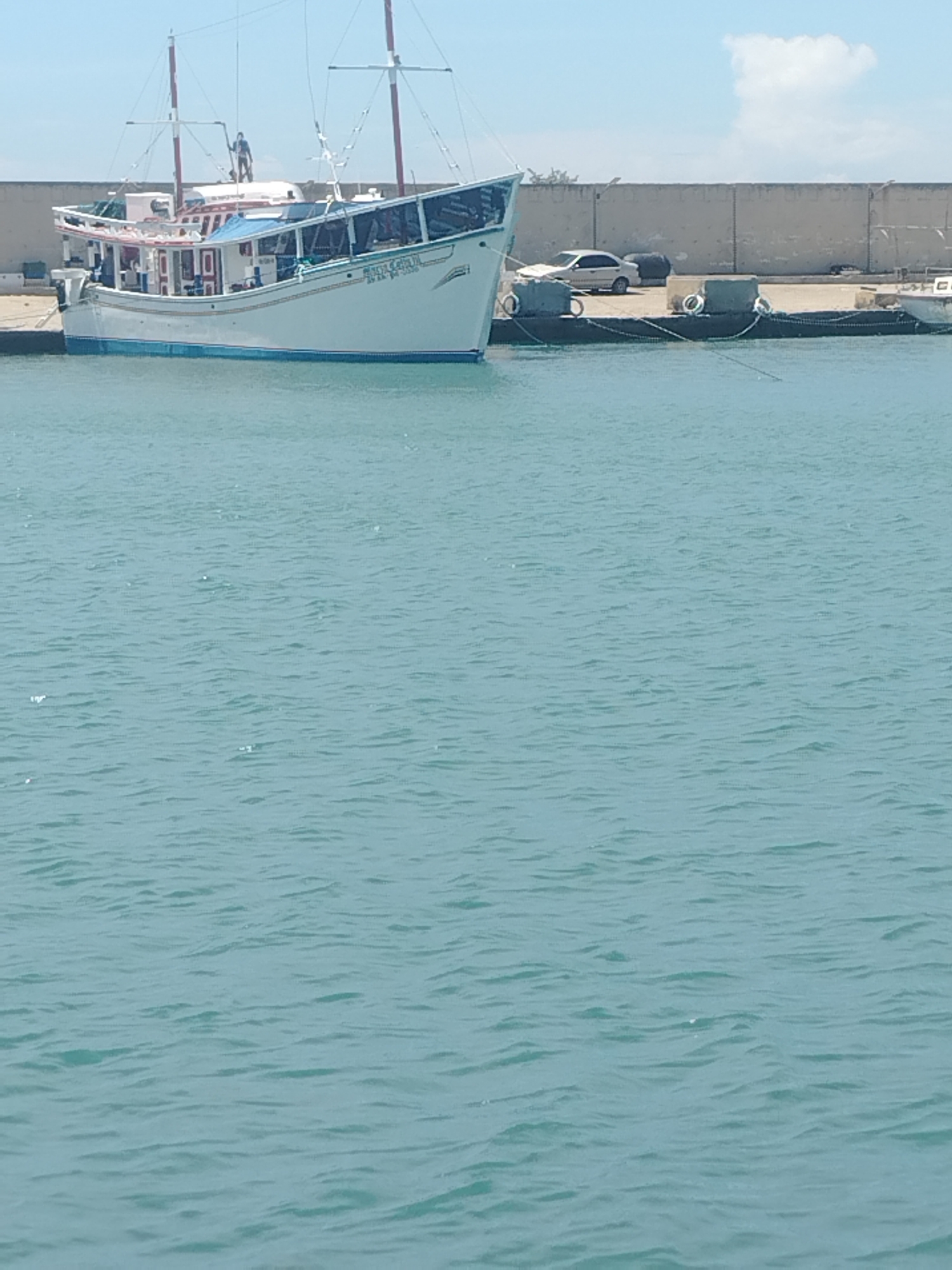 The media file depicts a daytime scene at a harbor in Porlamar, Venezuela. The primary subject is a white boat with blue trim, moored alongside a concrete pier. The boat has multiple masts and appears to be a fishing or passenger vessel, with windows and railings visible along its structure. A person is standing on the deck of the boat, near the front.
The pier is lined with a concrete wall and has several objects on it, including two dark, cylindrical buoys and a white sedan. Ropes are securing the boat to the pier. The water in the foreground is a tranquil blue, with small ripples on its surface, suggesting calm weather. The sky is a clear blue with a few white clouds visible.
The overall impression is one of a typical day at a harbor, with vessels and vehicles present, and the activity appearing to be routine docking or mooring. There is no overt indication of any specific event or activity beyond the presence of the boat and the parked car. The bright daylight suggests it is likely midday or early afternoon.