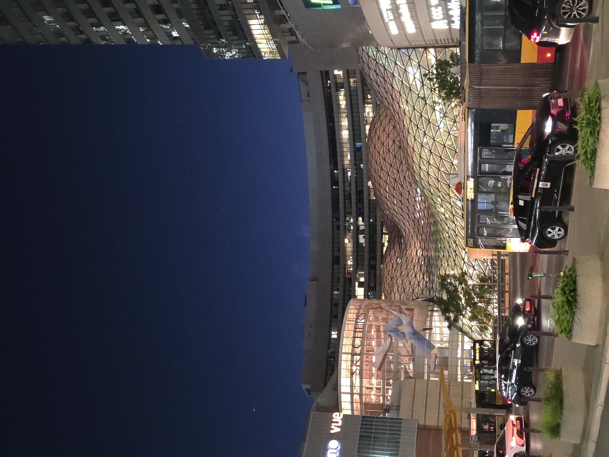 The image shows a nighttime cityscape in Warsaw, Poland. Modern buildings dominate the scene, with illuminated glass facades and distinctive architectural designs. A curved, lattice-like structure is a prominent feature, along with a building displaying a large advertisement or mural. Street-level activity is visible, with parked cars and buses lined along the curb. The sky is dark blue, suggesting a late evening or night setting.