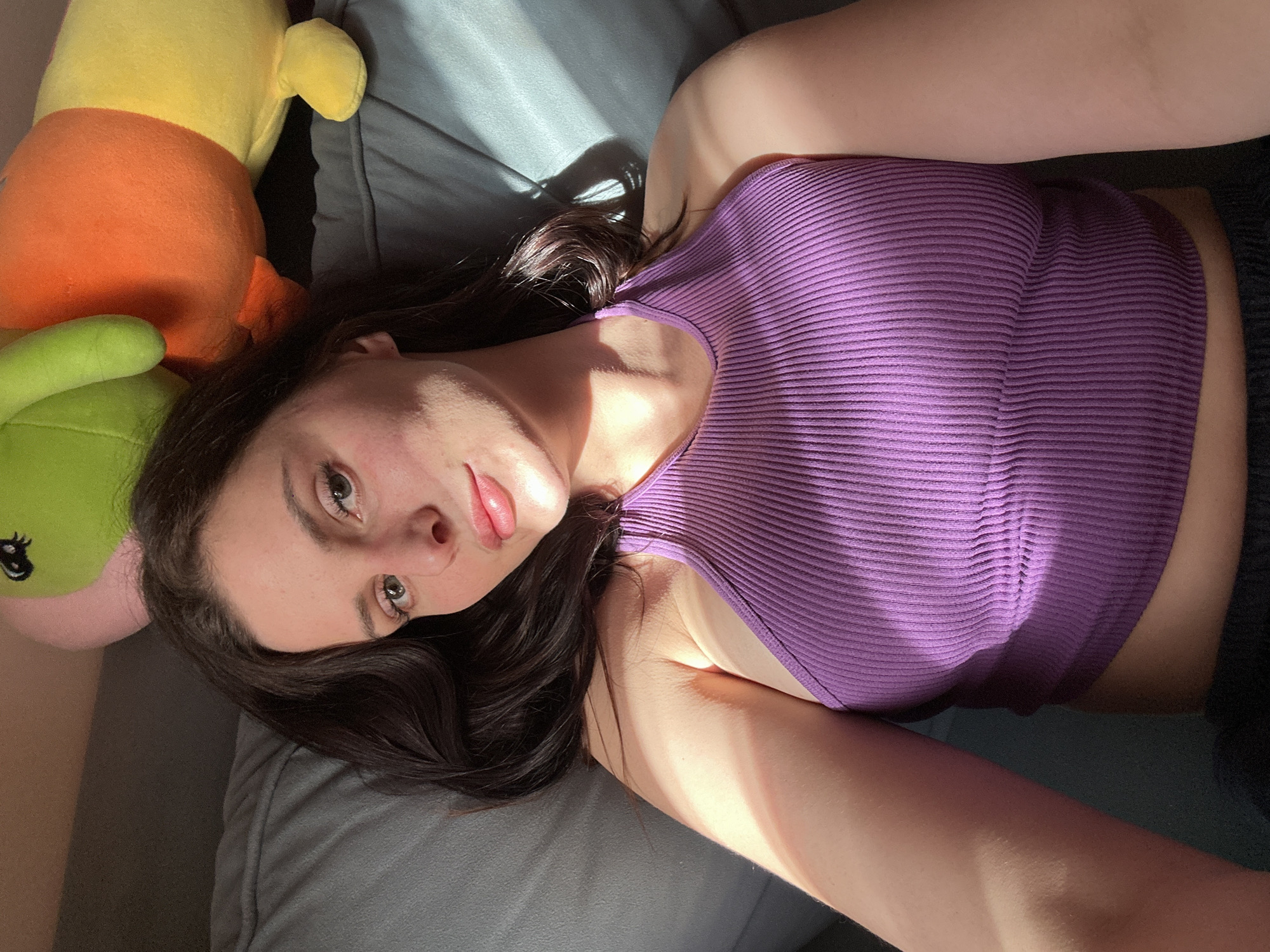 The media file is a close-up, high-angle, portrait-oriented photograph of a young woman lying on her side on a couch. She is wearing a ribbed, purple tank top. Her head is propped up, and her gaze is directed towards the camera. Sunlight casts strong shadows across the scene, suggesting it is daytime, likely late afternoon or early morning. To her left are parts of two stuffed animals, one yellow and orange, and another green. The overall mood appears relaxed and casual. The background is dark, with shadows obscuring details of the couch and surrounding environment. There is no visible text in the image.