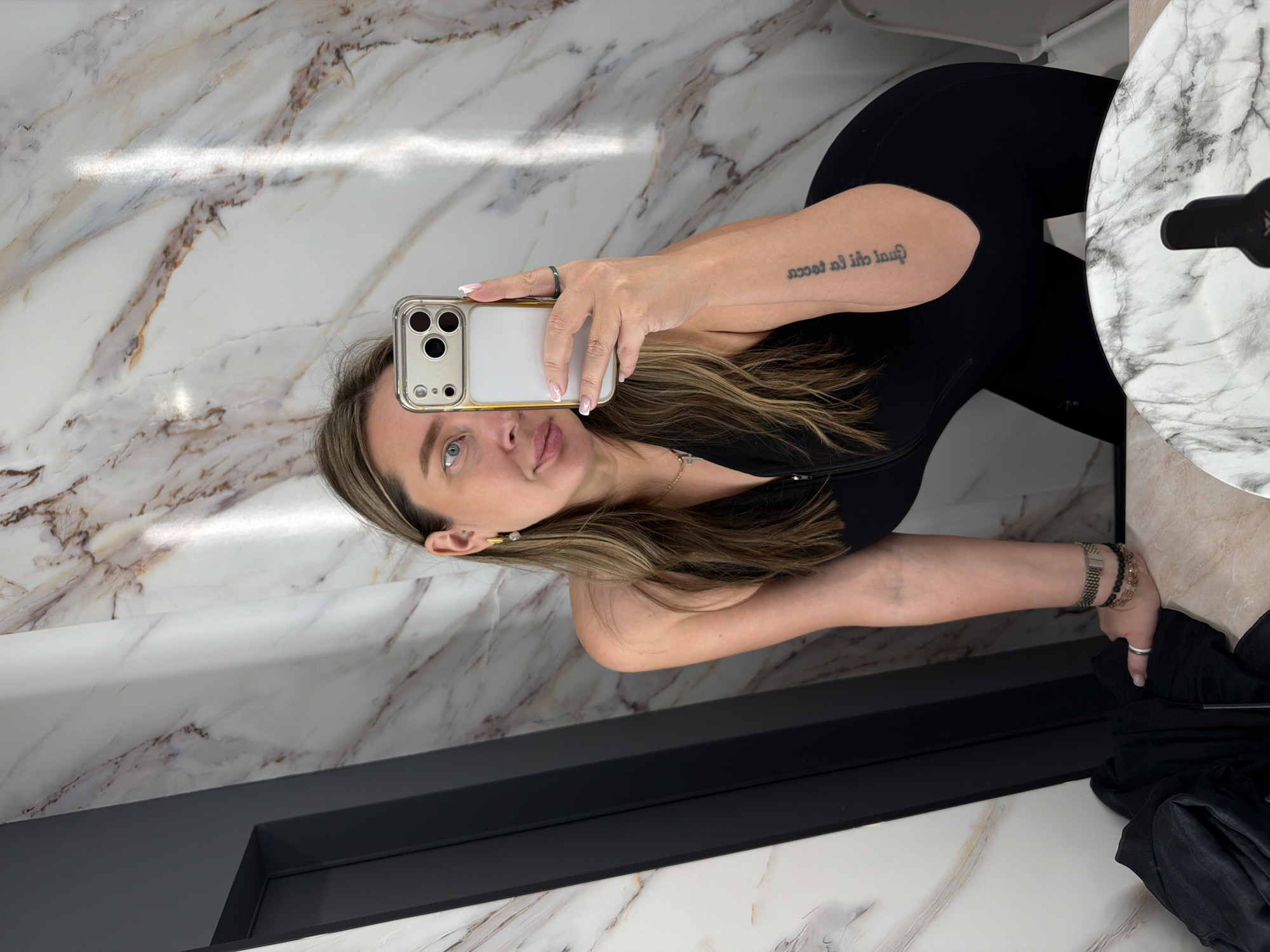 A woman is taking a selfie in what appears to be a bathroom. The bathroom has a marble wall and countertop. The woman is wearing black athletic clothing. She is holding a gold iPhone in her left hand, and the reflection shows her face. She has shoulder-length brown and blonde hair, blue eyes, and a fair complexion. She is wearing a ring on her left hand, several bracelets on her right wrist, and a gold necklace. There is a tattoo on her left arm. A black object is visible to the right of the counter. The location is not recognizable as Moscow, Russia from the available information.