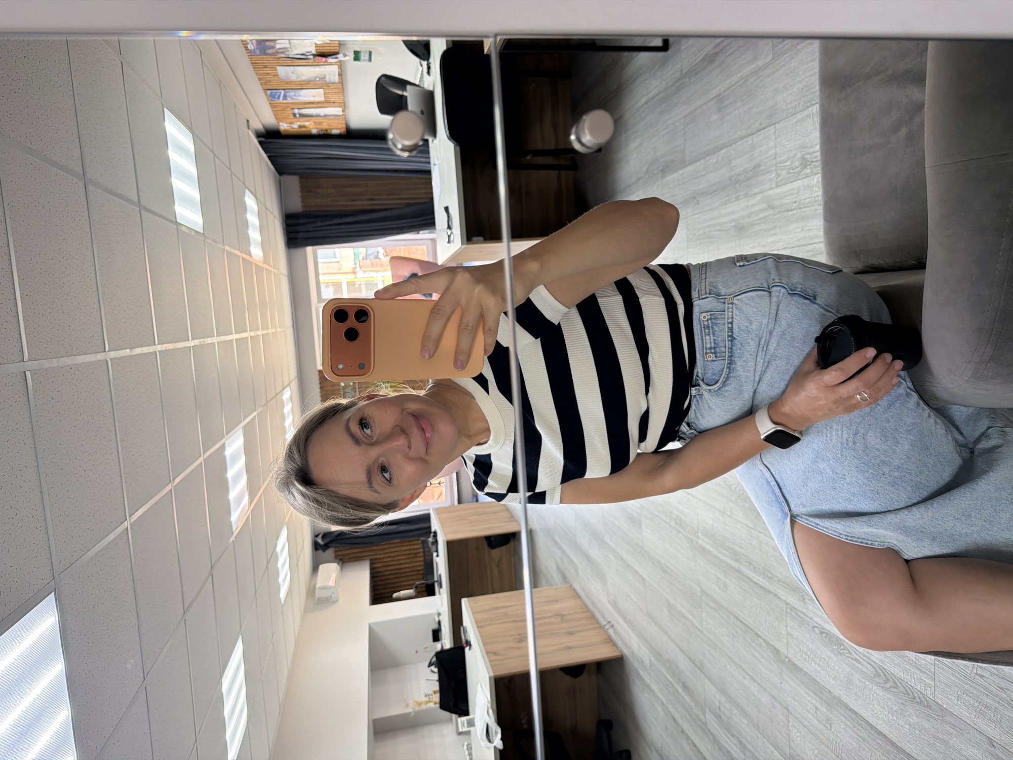 The media shows a vertical, mirrored selfie taken in an indoor setting. The primary subject is a woman with blonde hair, wearing a black and white striped top and light blue denim shorts. She is holding a smartphone in her right hand, angled to capture her reflection. Her left arm is resting on a light grey couch, and a black camera is visible in her left hand. She is wearing a smartwatch on her left wrist and a ring on her right hand.
The setting appears to be a modern office or studio space, reflected in a large mirror. The ceiling is covered with acoustic tiles and features multiple rectangular fluorescent light fixtures. The floor is a light grey wood laminate. In the background reflections, we can see wooden desks, office chairs, and what looks like a window with natural light coming through. The overall lighting suggests it is daytime. The woman's facial expression is a slight smile, conveying a casual and relaxed mood. There is no visible text in the image.