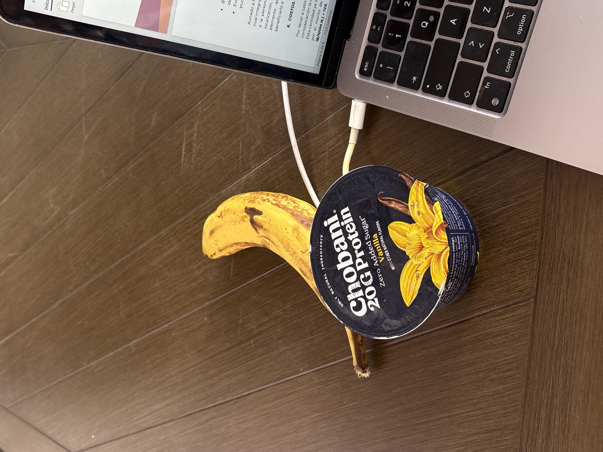 The image presents an overhead view of an indoor setting, possibly a wooden floor or table, where a silver laptop and snacks are arranged. A laptop, resembling a MacBook, is positioned on the right side of the frame, with its screen displaying a document in Spanish. A white charging cable is plugged into the laptop.
Beside the laptop, a Chobani Zero Sugar Vanilla yogurt cup is lying on its side, revealing its label. The label clearly states "Chobani. 20G Protein Zero Added Sugar* Vanilla WITH OTHER NATURAL FLAVORS," along with nutritional details like "Total Fat 3g" and "Protein 20g." A ripe, yellow banana is placed next to the yogurt cup.
The laptop screen shows fragments of a document, including headers like "6. COSTOS." and other Spanish text such as "Página 5 de 6" and "1123 palabras," indicating a work or study activity. The laptop's keyboard, showing keys like "esc," "control," and "option," is also partially visible.
The scene suggests a moment of leisure or a break from work, with healthy snacks accompanying the digital activity. There are no individuals present, and the indoor environment does not offer specific cues about the time of day or weather. The Spanish text on the laptop is consistent with the given location context of Puebla, Mexico.