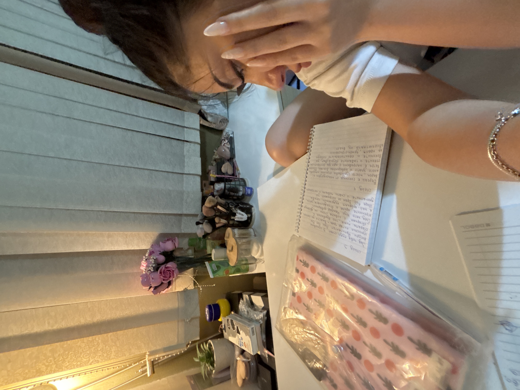 In Plovdiv, Bulgaria, a young woman with dark hair tied up is seated at a desk, appearing to be studying or working. She is wearing a white t-shirt and has long, light-colored fingernails. One hand is resting on her face, near her eye. The desk is cluttered with various items, including an open notebook with handwritten text, a plastic-wrapped package containing what appears to be a pink and white patterned item with small dark shapes, and a collection of makeup brushes in a holder on the right side of the desk. Behind the desk, there are vertical blinds on a window, and to the left, a bouquet of pink flowers in a vase is visible, along with a small potted plant and various cosmetic products and containers. The lighting suggests it is daytime or evening with artificial light. The scene appears to be an indoor study or personal space.